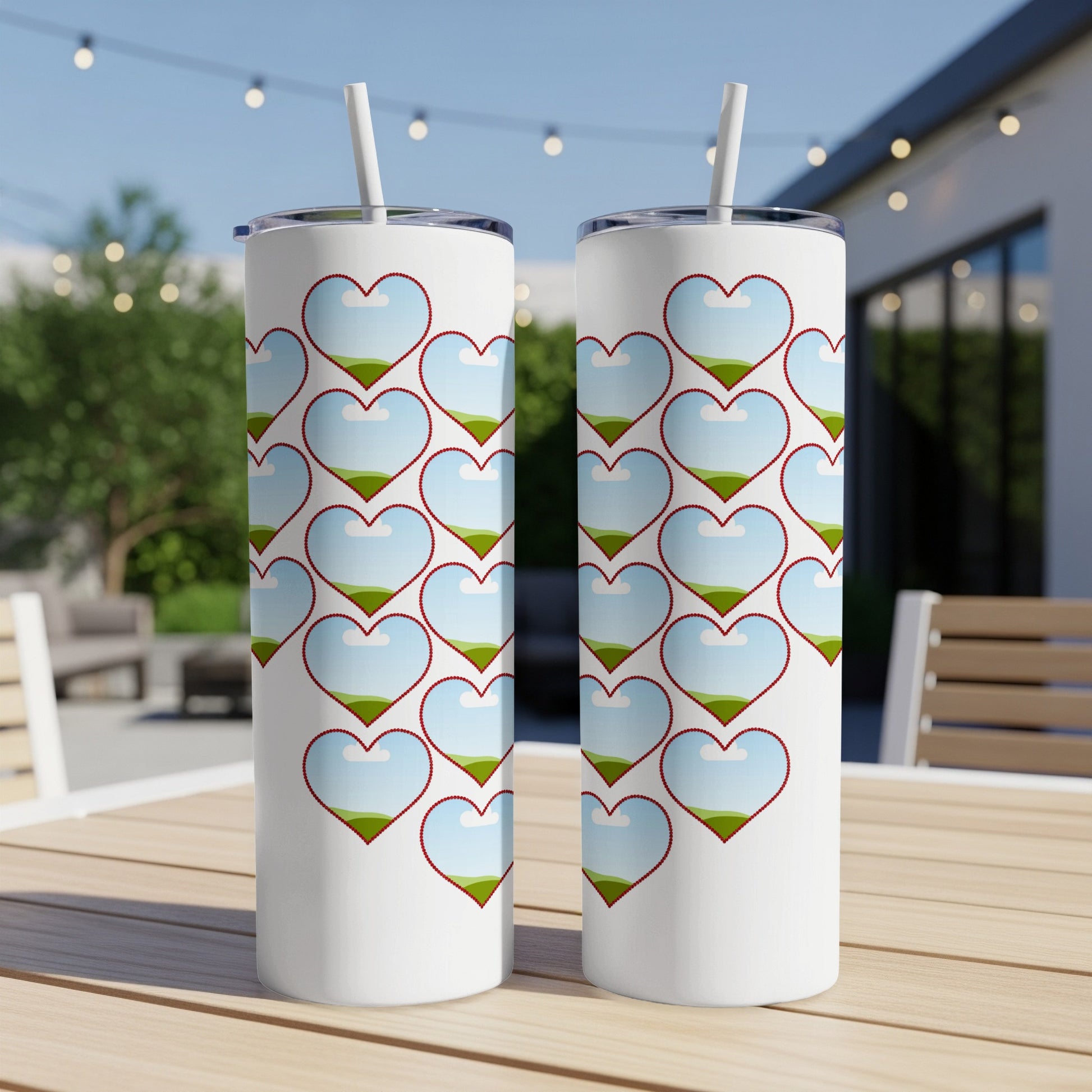 Two white tumblers with a repeating pattern of red-bordered heart shapes filled with a landscape of sky, clouds, and green hills.