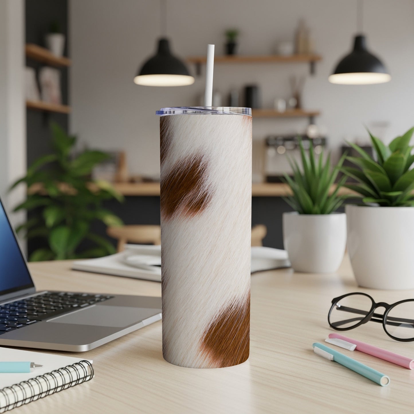Cow print skinny tumbler with straw on a desk with laptop and pens.