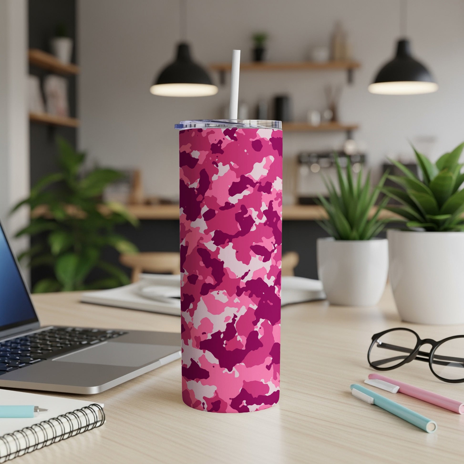 Pink camouflage tumbler with straw on wooden desk next to laptop and notebook.