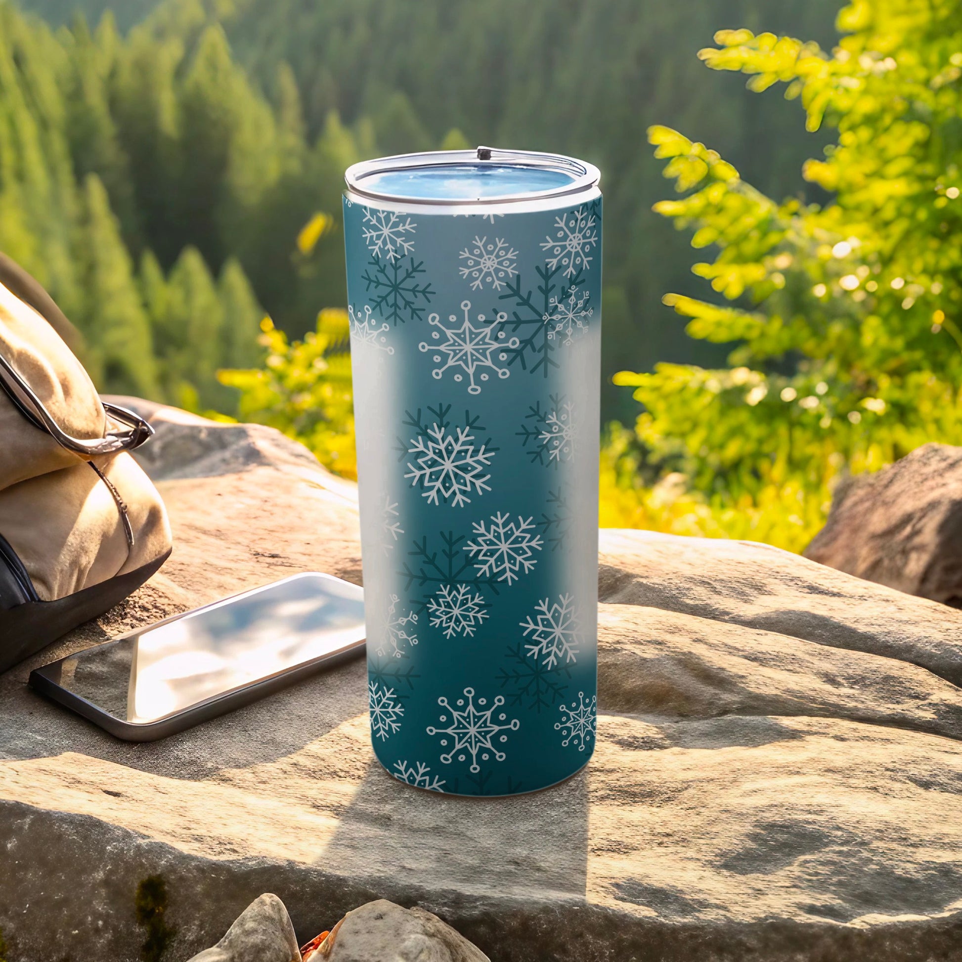 Teal tumbler with white snowflakes, next to phone and backpack.