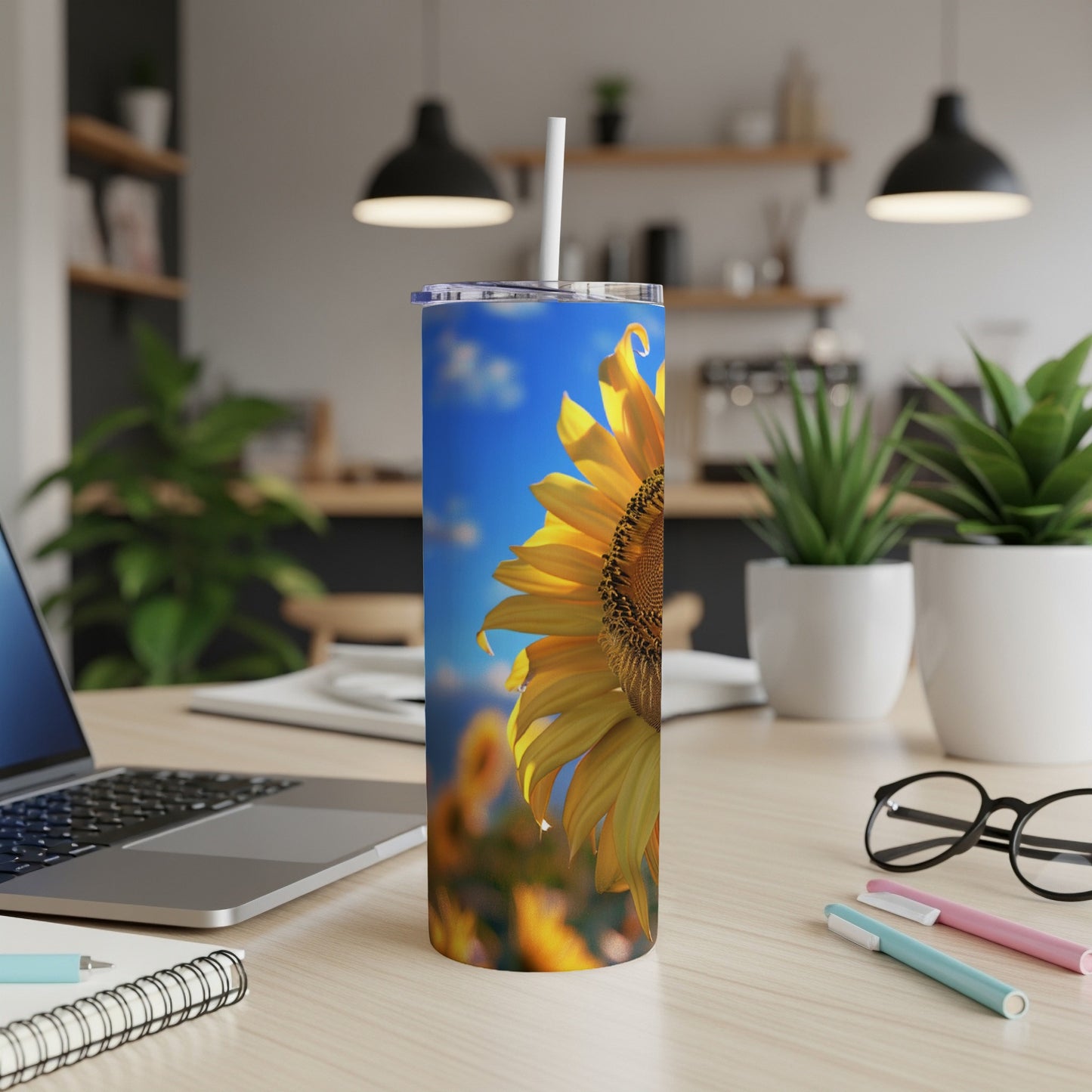 Sunflower on a tumbler with a straw, laptop, and pens on a desk.