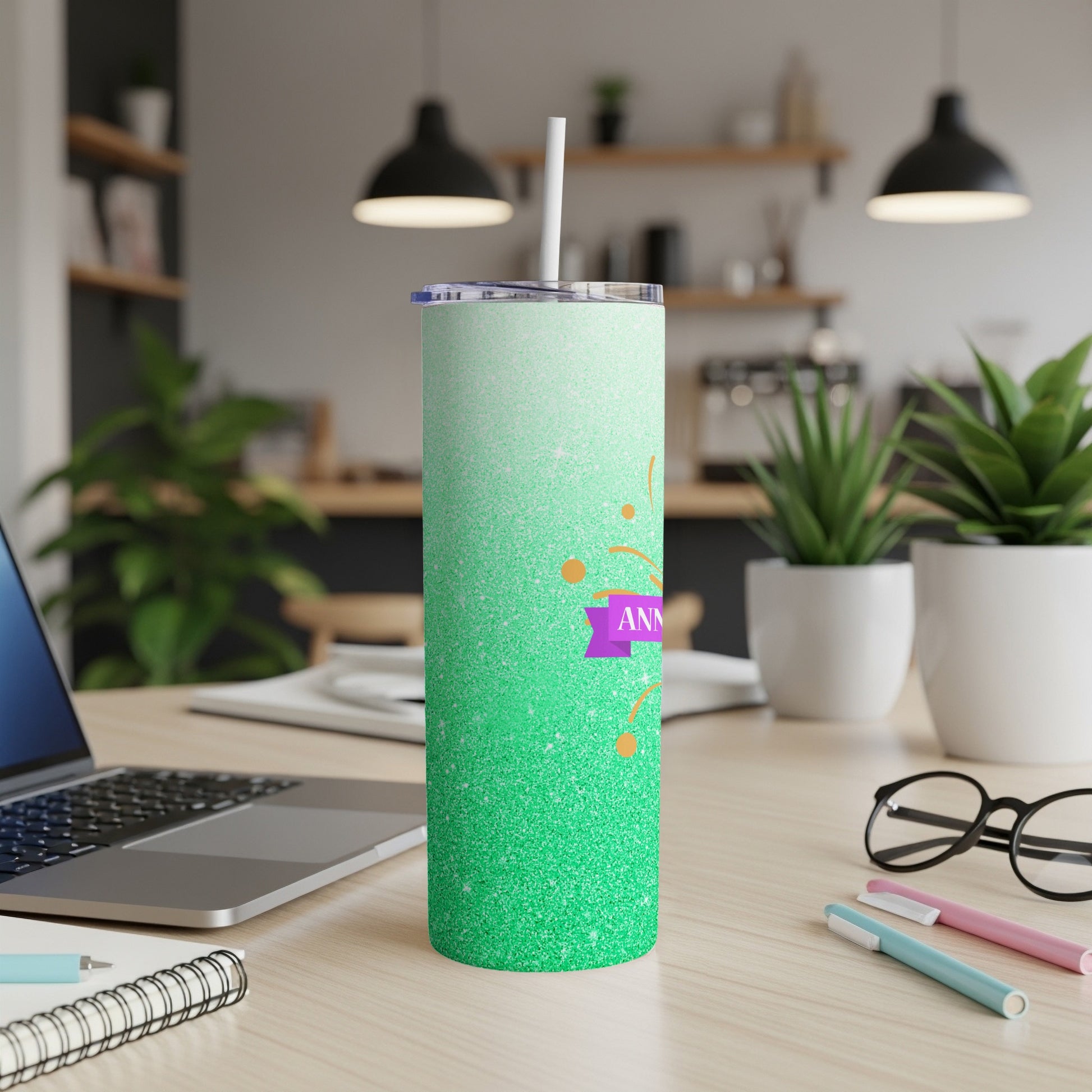 Green glitter tumbler with "ANNIVERSARY" ribbon and straw on desk.
