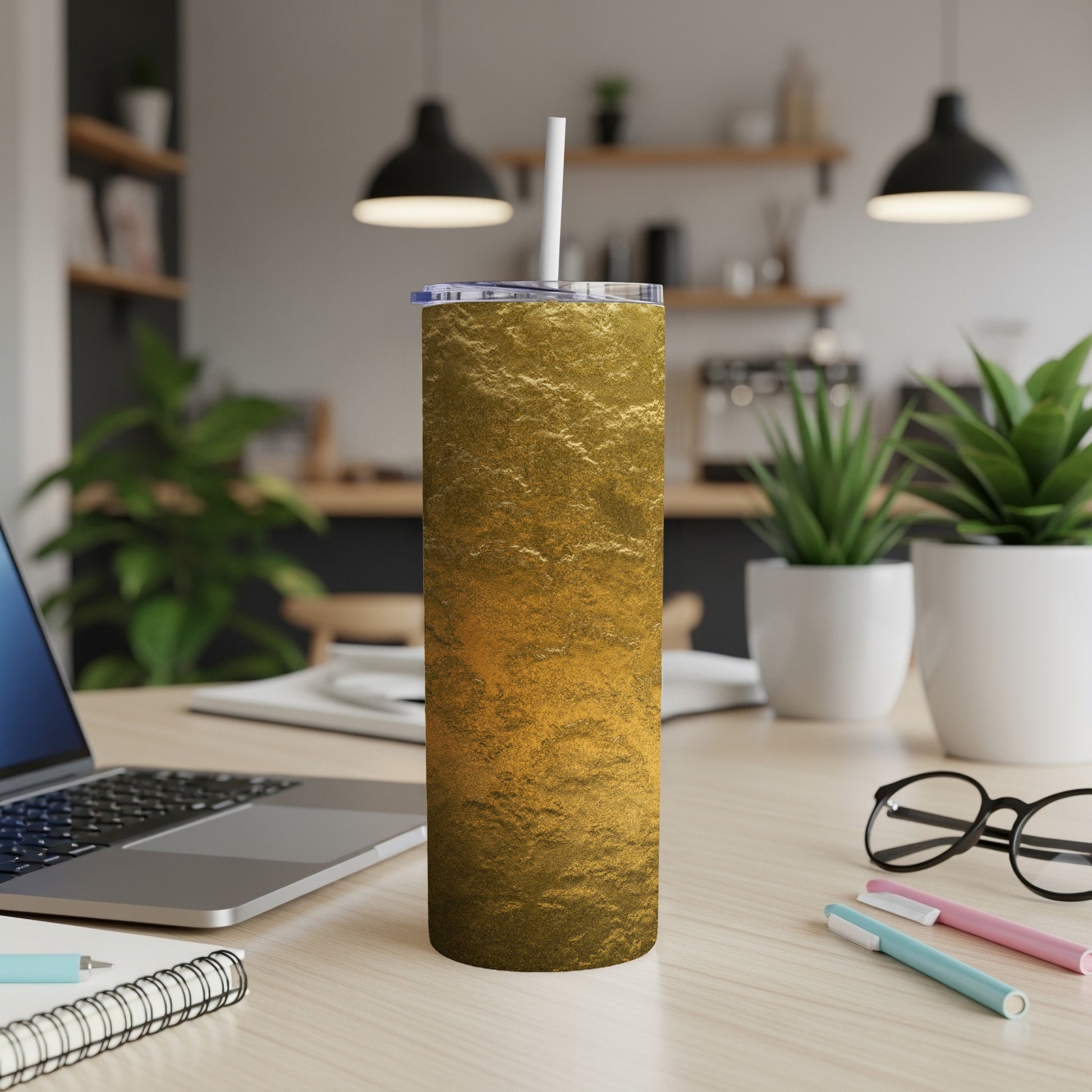 Gold textured tumbler with white straw on a wooden desk with laptop and plants.