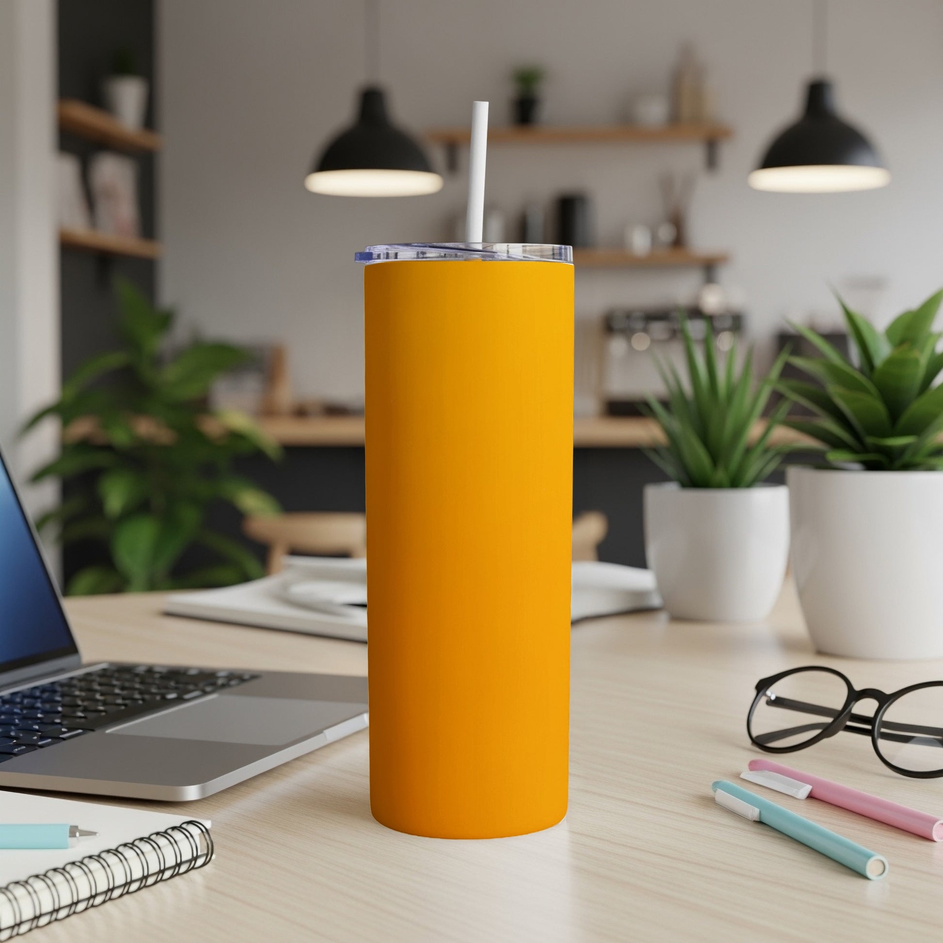 Orange tumbler with straw on a desk next to a laptop and pens.
