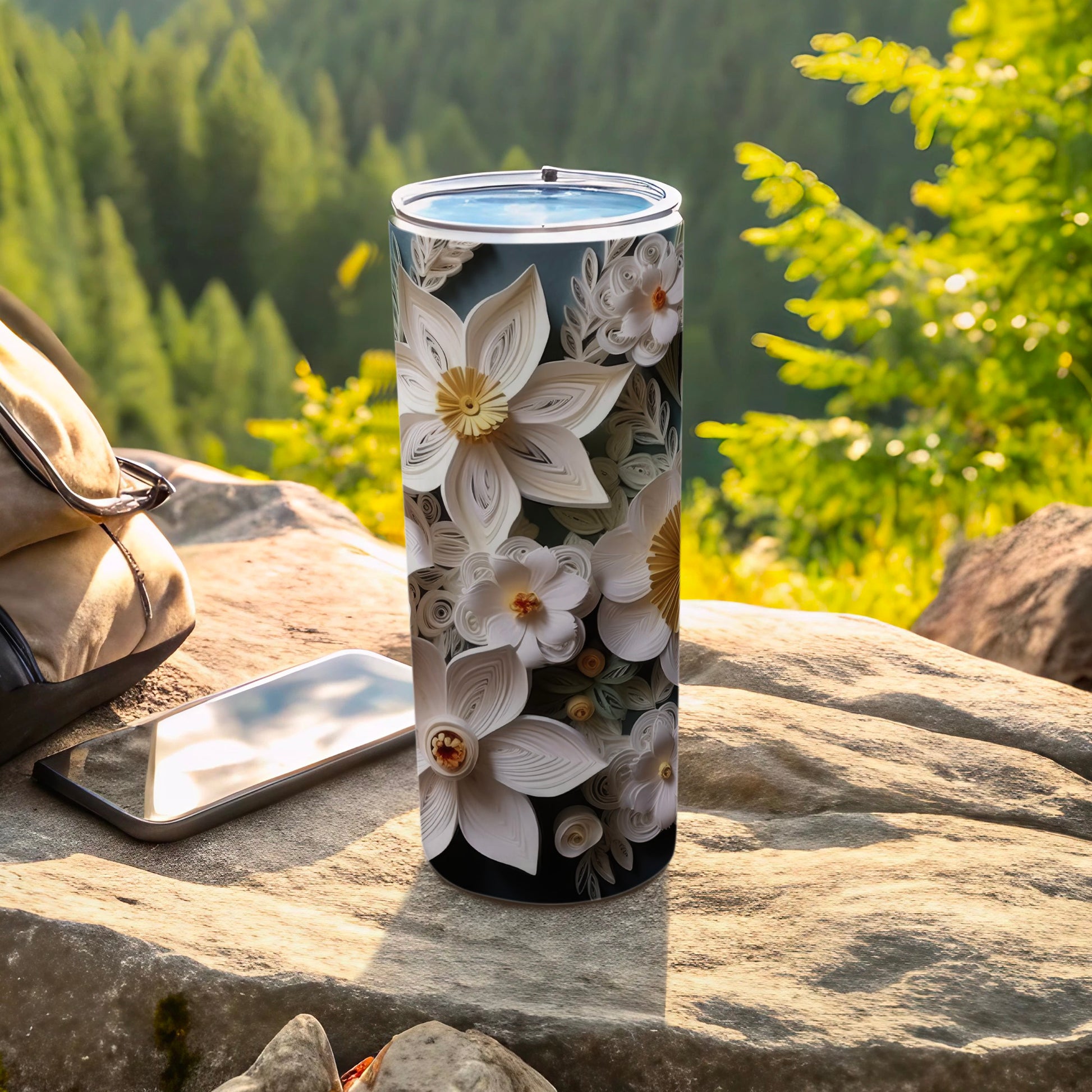 Floral tumbler on rocks with backpack and phone, sunny forest background.
