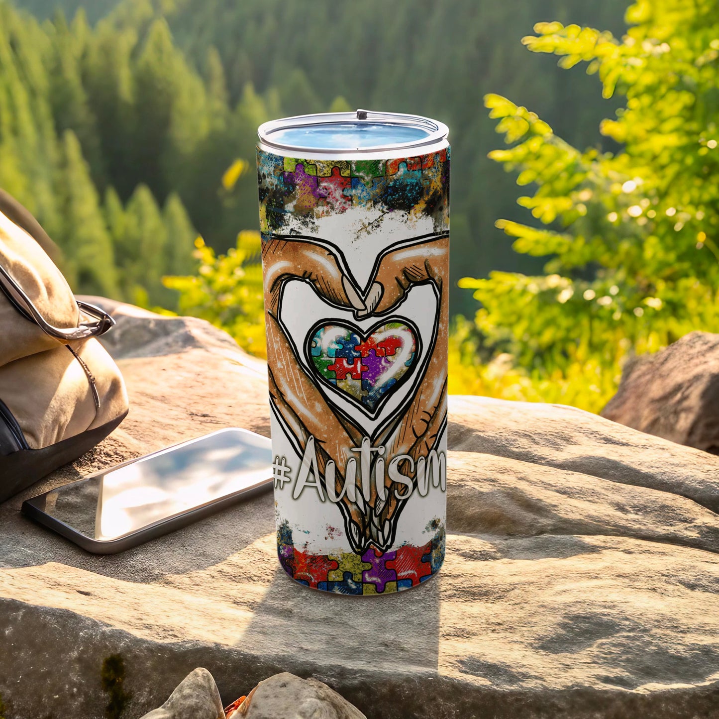 Autism puzzle piece heart tumbler with hands, on a rock outdoors.