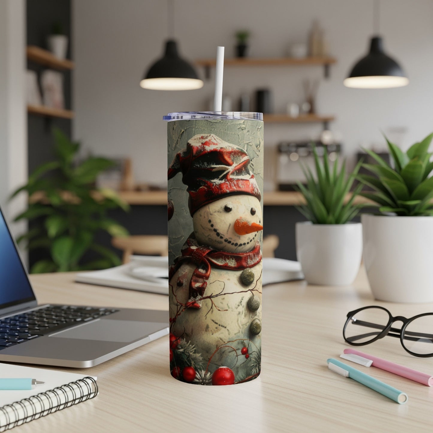 Snowman tumbler with red scarf and hat on desk with laptop and pens.
