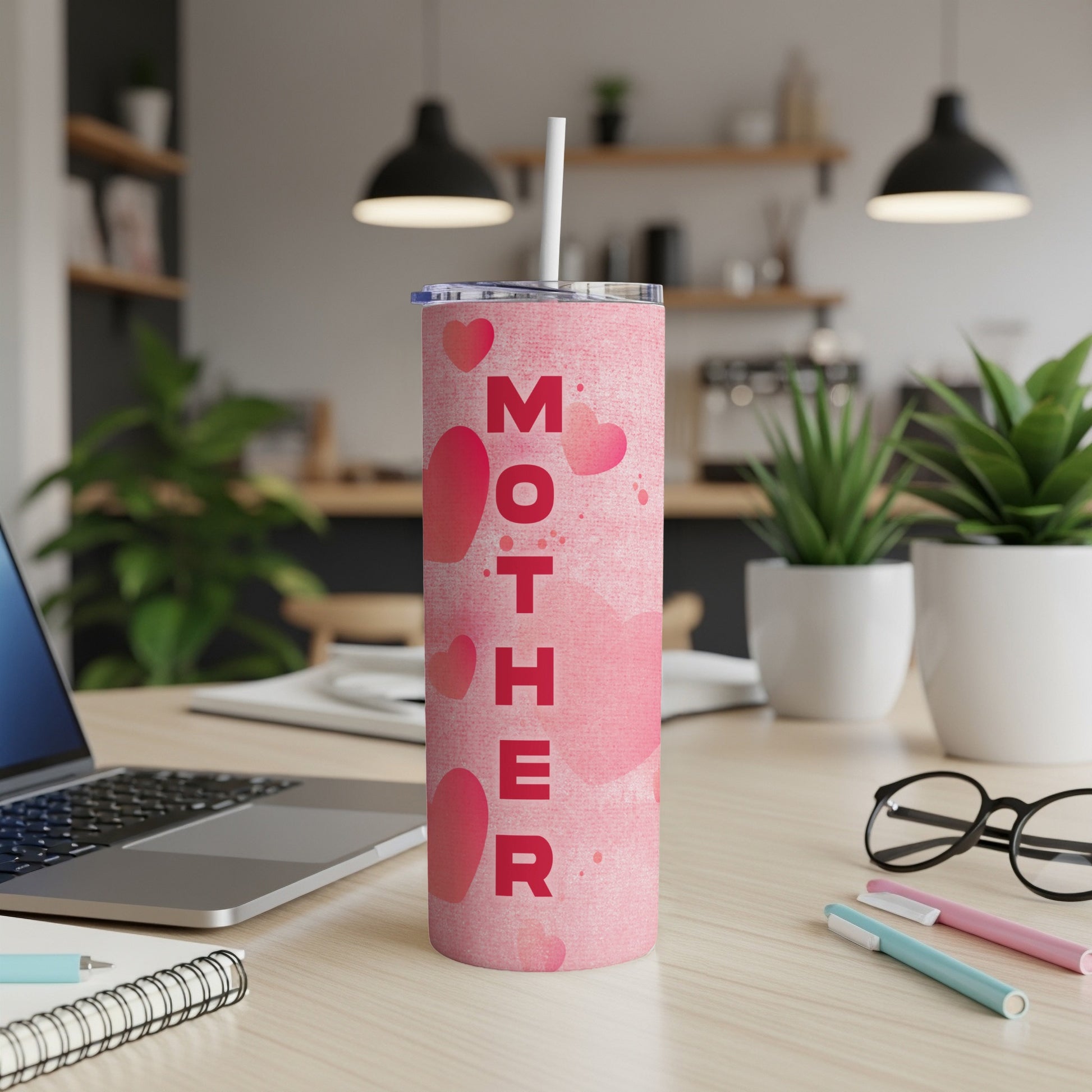 Pink tumbler with hearts and "MOTHER" text on a desk with a laptop.