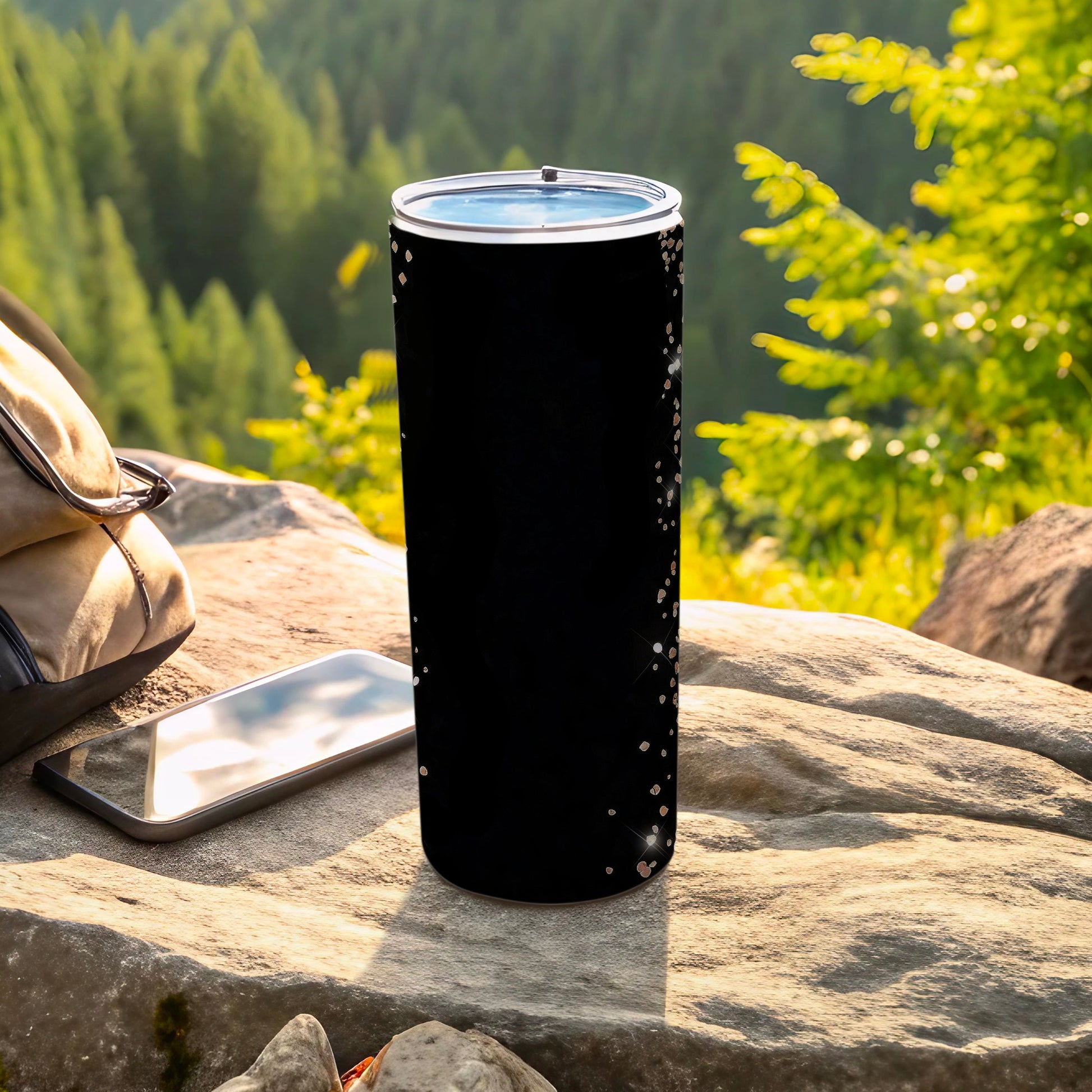Black tumbler with glitter speckles, resting on rock with phone and bag.