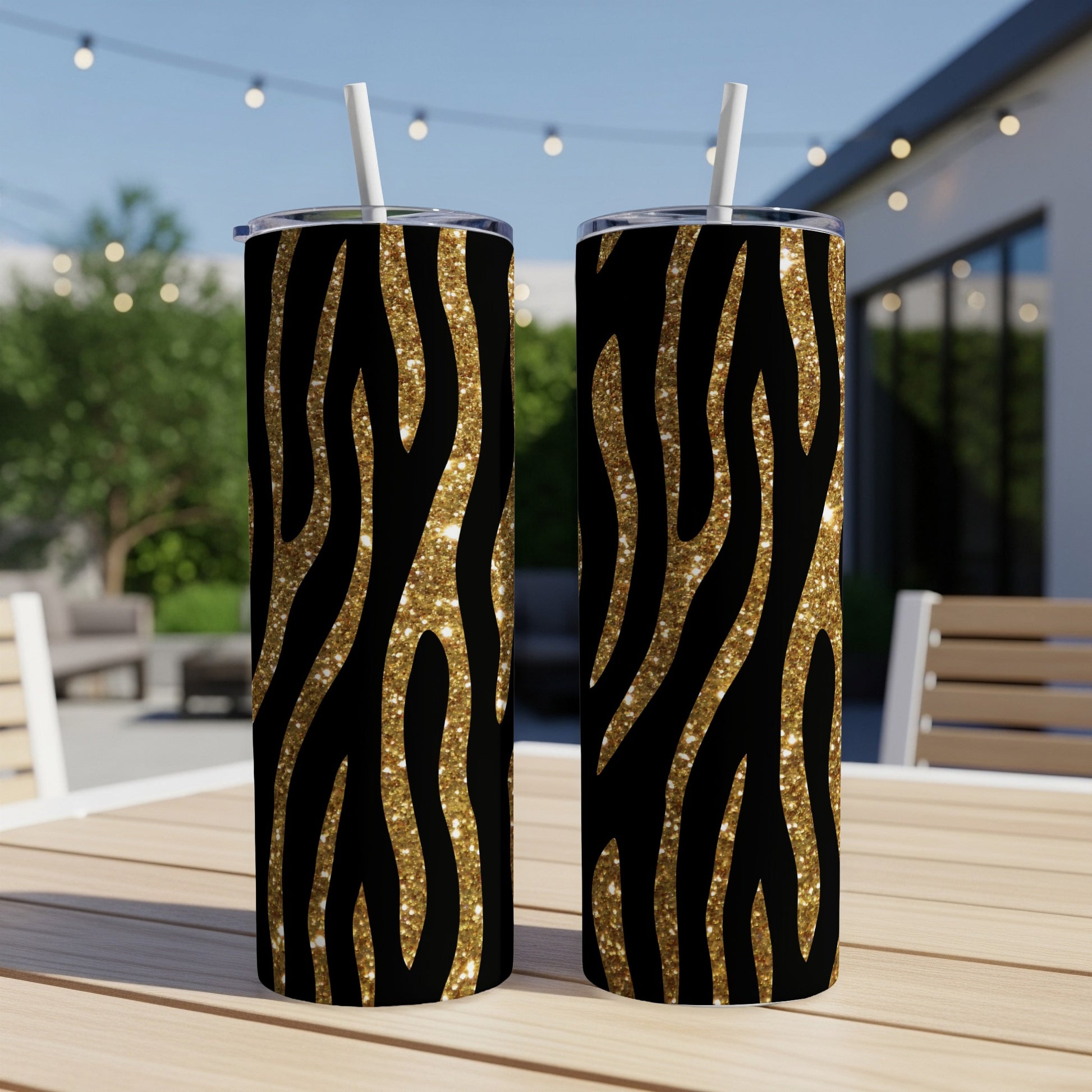 Two tumblers with black and gold glitter zebra print and white straws displayed outdoors.