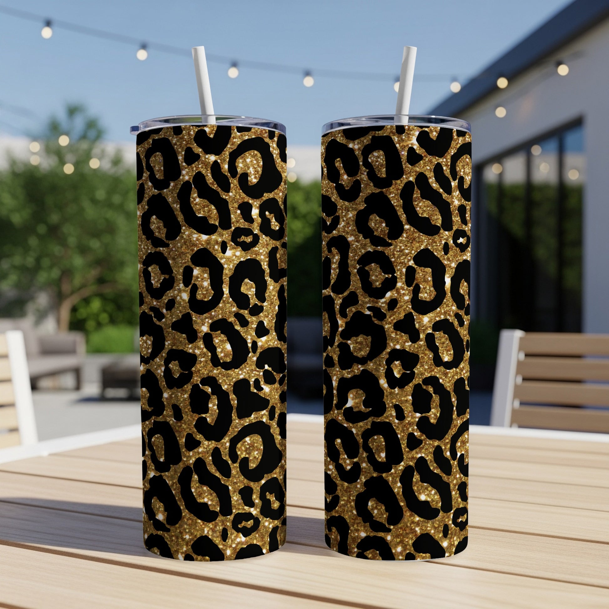 Two gold glitter leopard print tumblers with white straws on a wooden table outdoors.