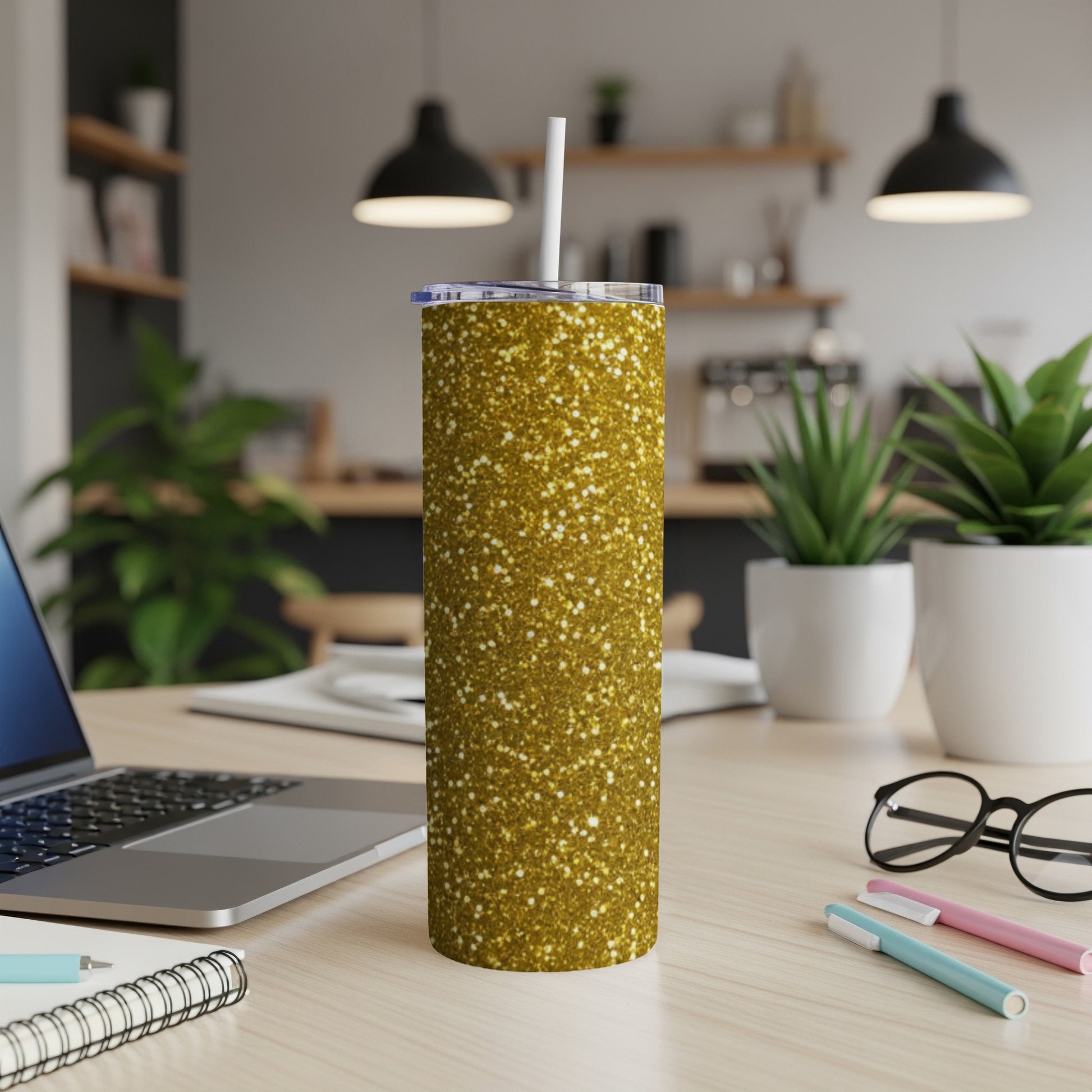 Gold glitter tumbler with straw on a wooden desk with laptop and plants.