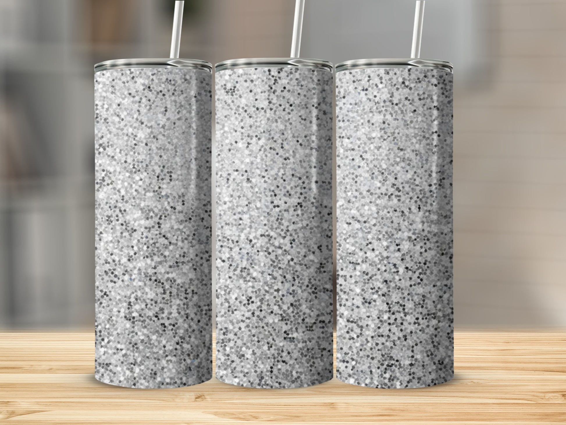 Three glittery silver tumblers with straws stand on a wooden surface.