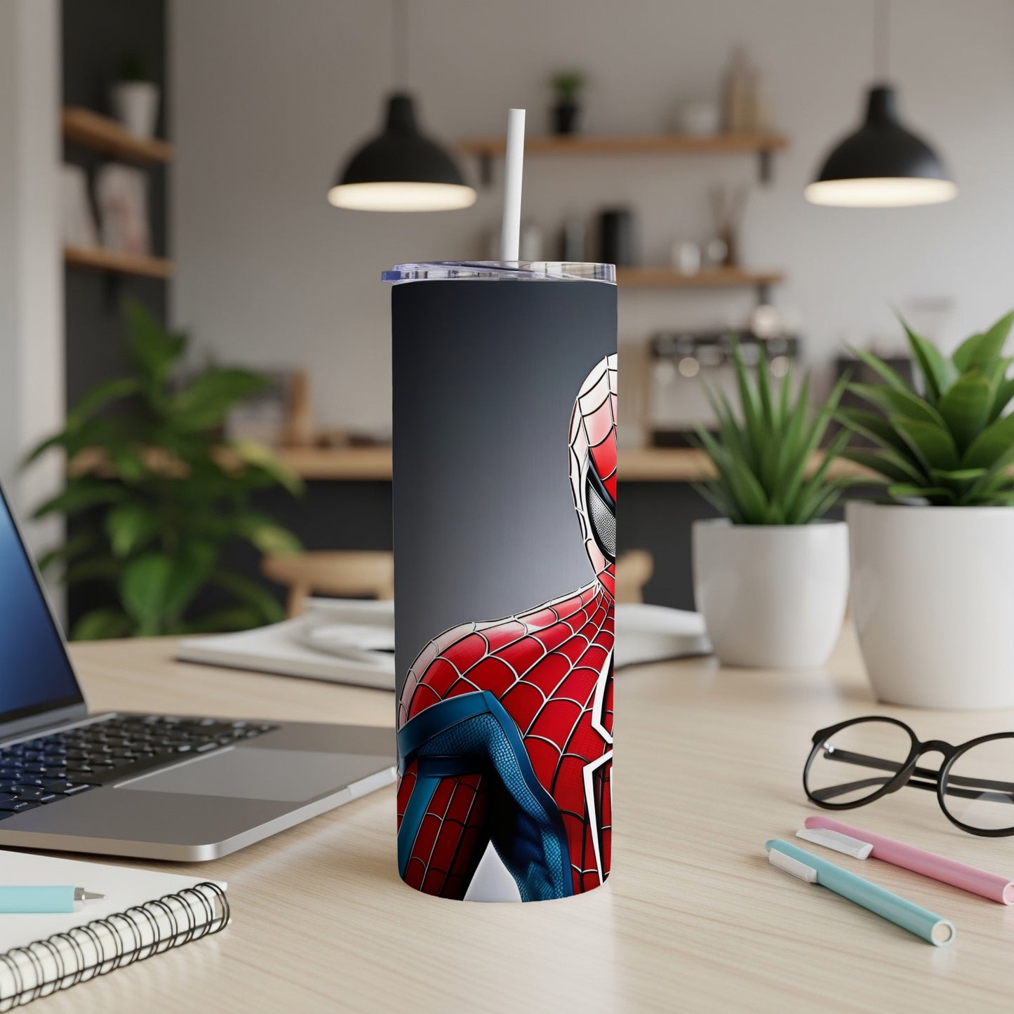 Spider-Man tumbler with straw on desk next to laptop and pens.
