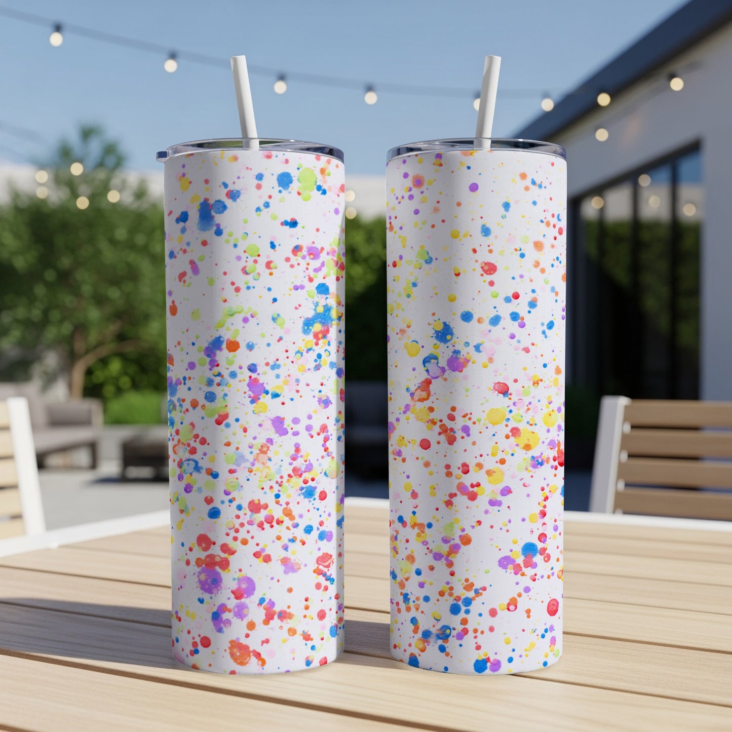 Two white tumblers with colorful paint splatter pattern, on a wooden table outdoors.