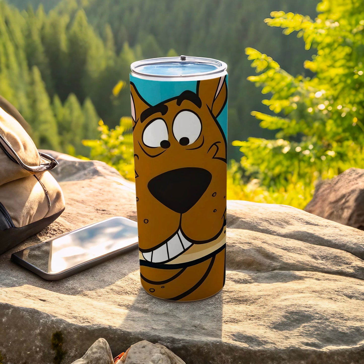 Scooby-Doo tumbler with character face on a rock, smartphone and backpack nearby.