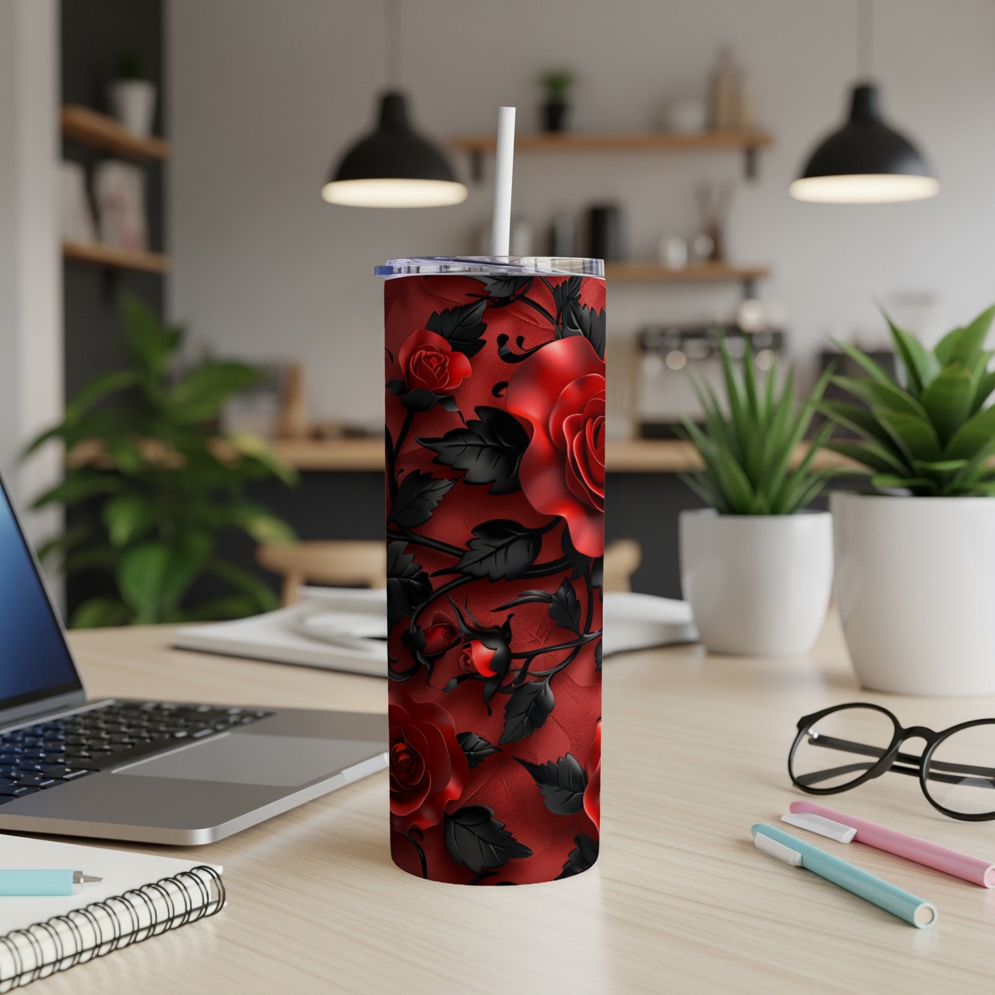 Red roses and black leaves adorn a white tumbler with a straw on a desk.