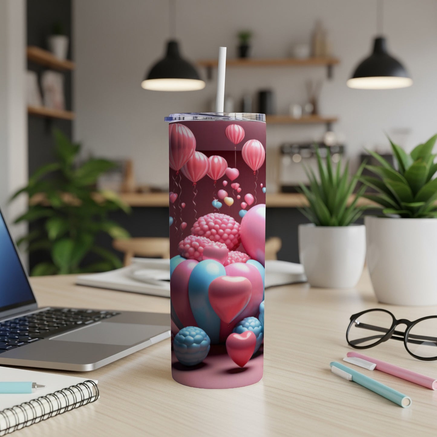 Pink tumbler with heart and balloon pattern, on desk with laptop.