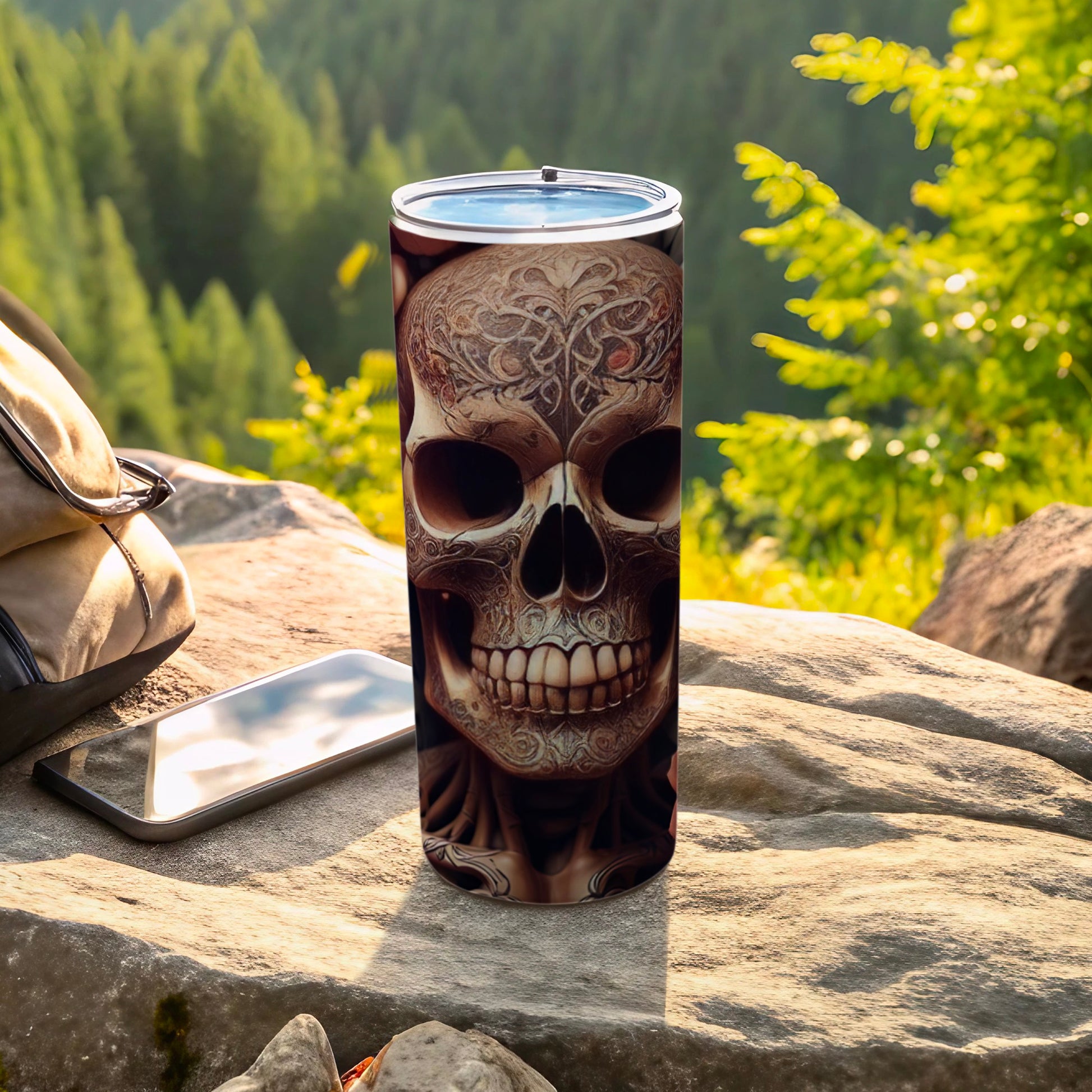 Tumbler with ornate skull design sits on rock with backpack and phone.