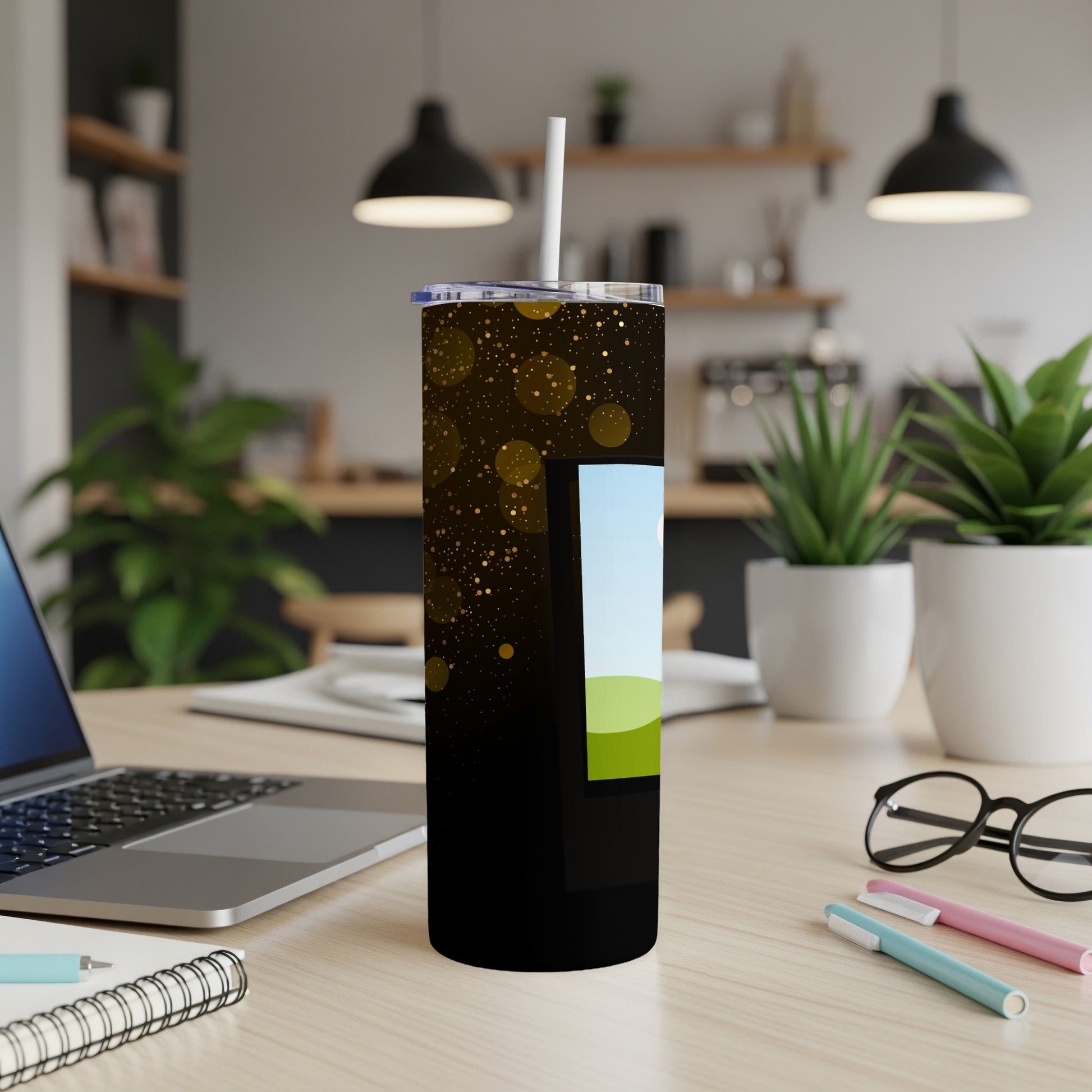 Black tumbler with gold glitter and a green landscape graphic, on a desk.