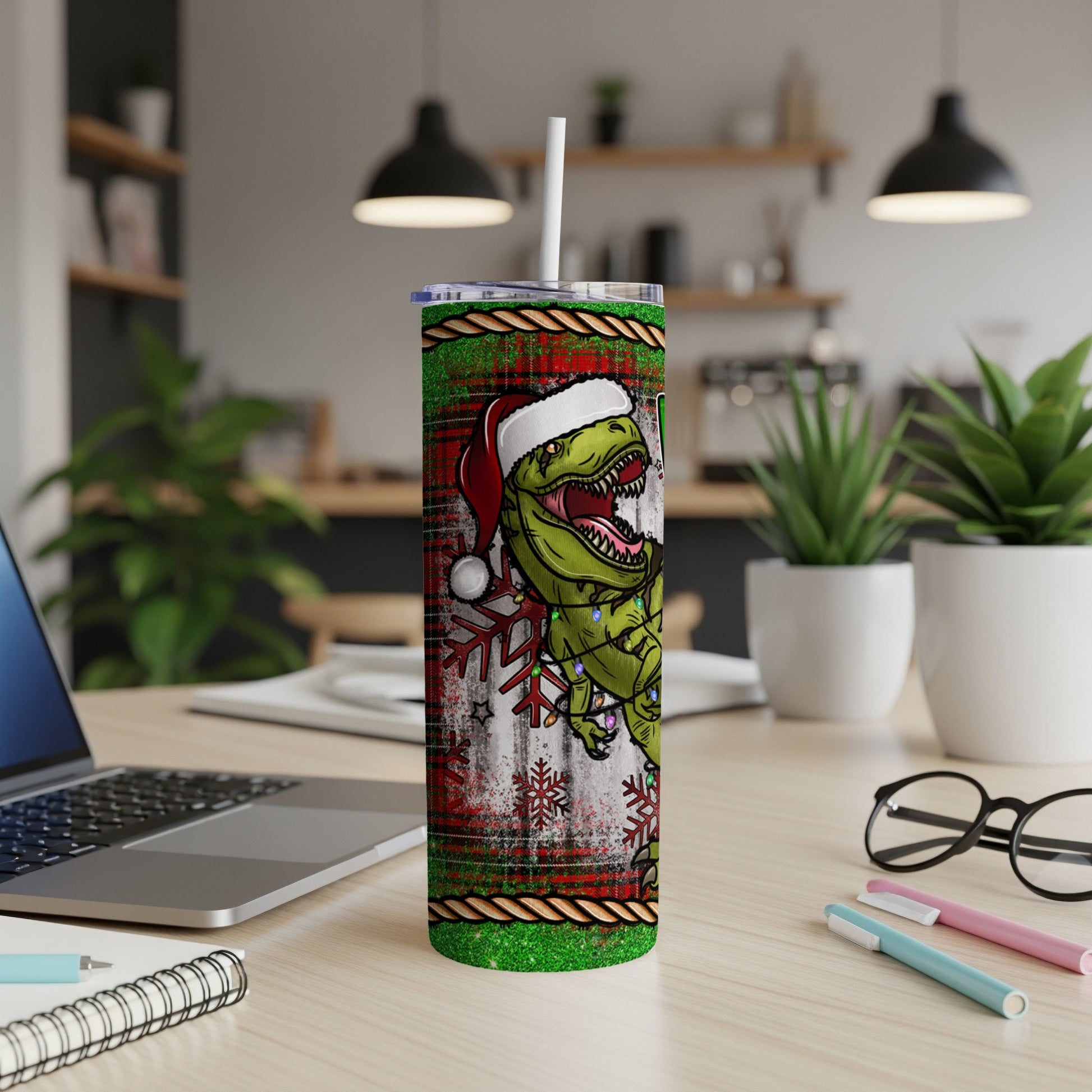 Christmas dinosaur tumbler with Santa hat, snowflakes, and lights on a desk.