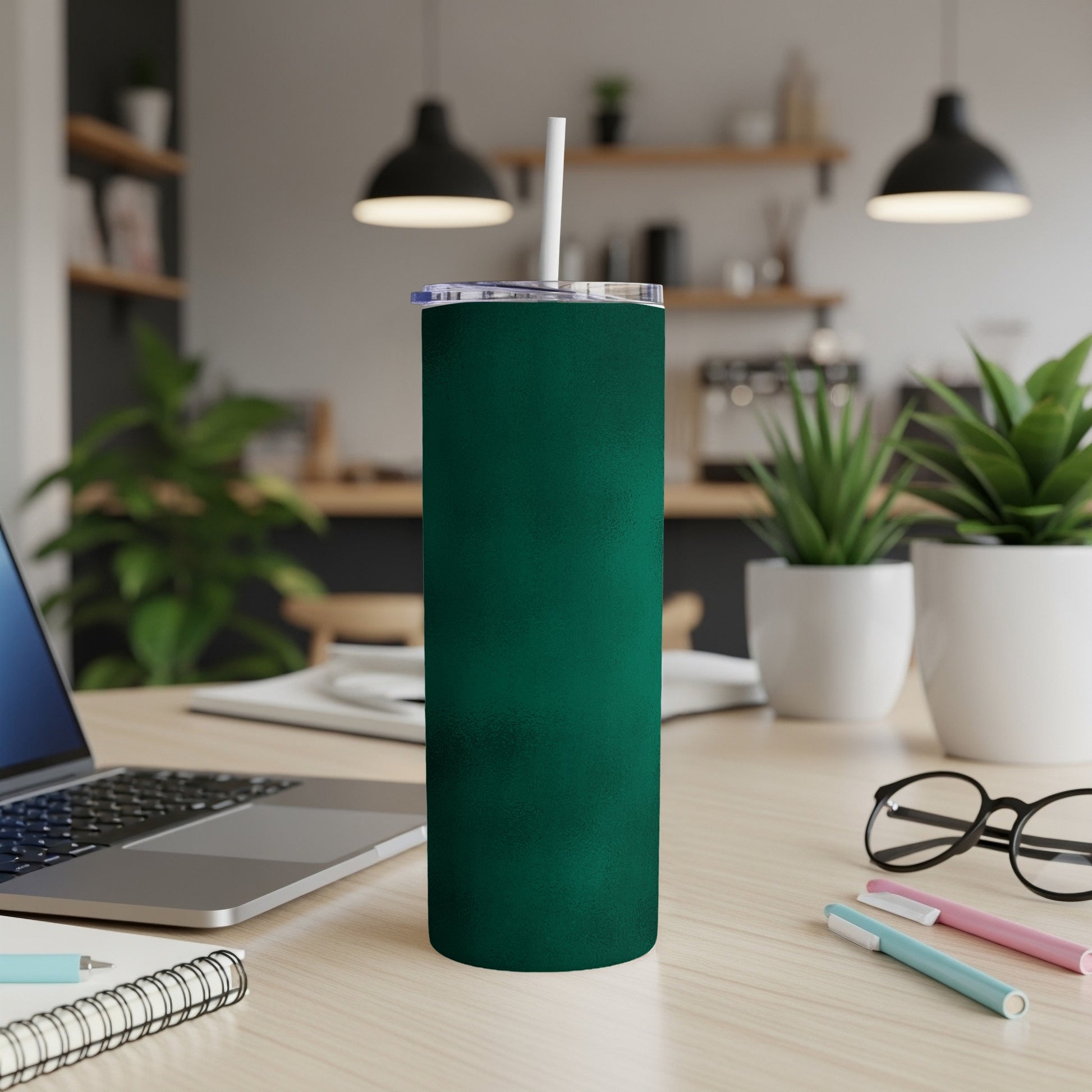 Dark green tumbler with white straw on a desk with laptop and plants.