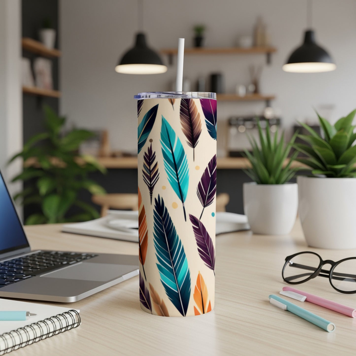 Feather-patterned tumbler with straw on a desk next to a laptop.