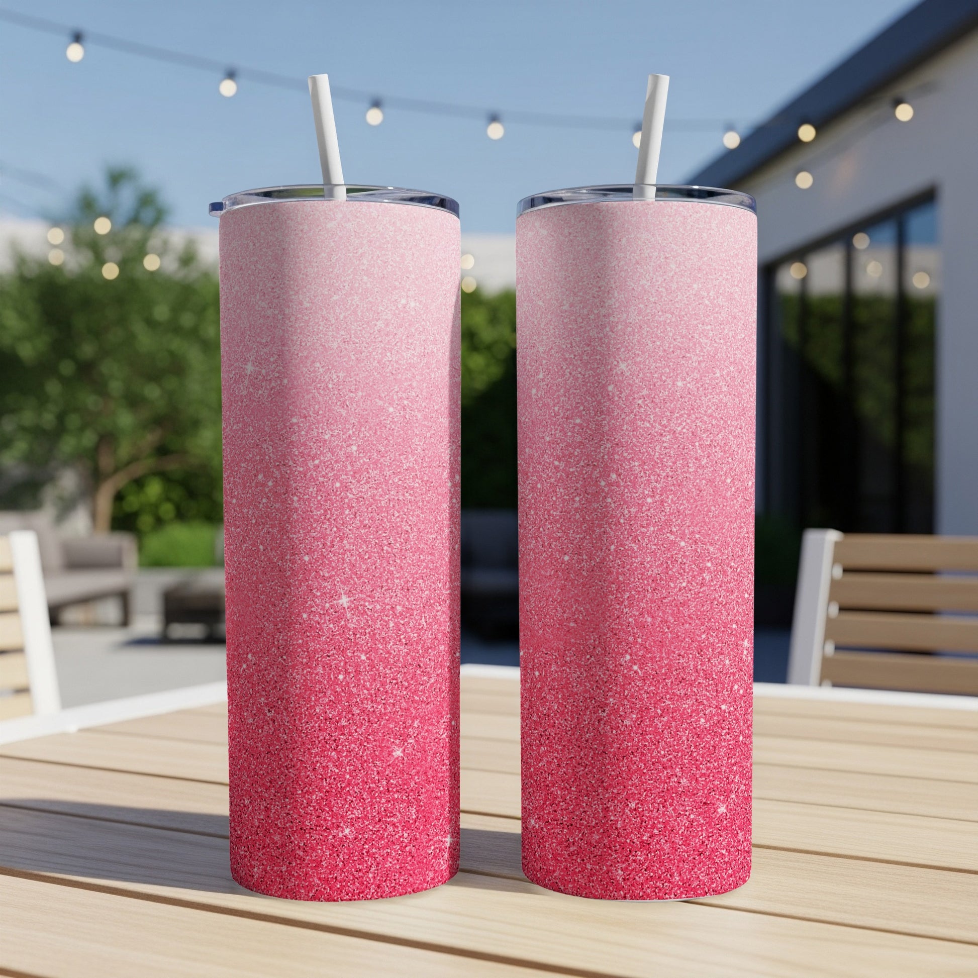 Two glittery pink ombre tumblers with straws on a table outdoors.