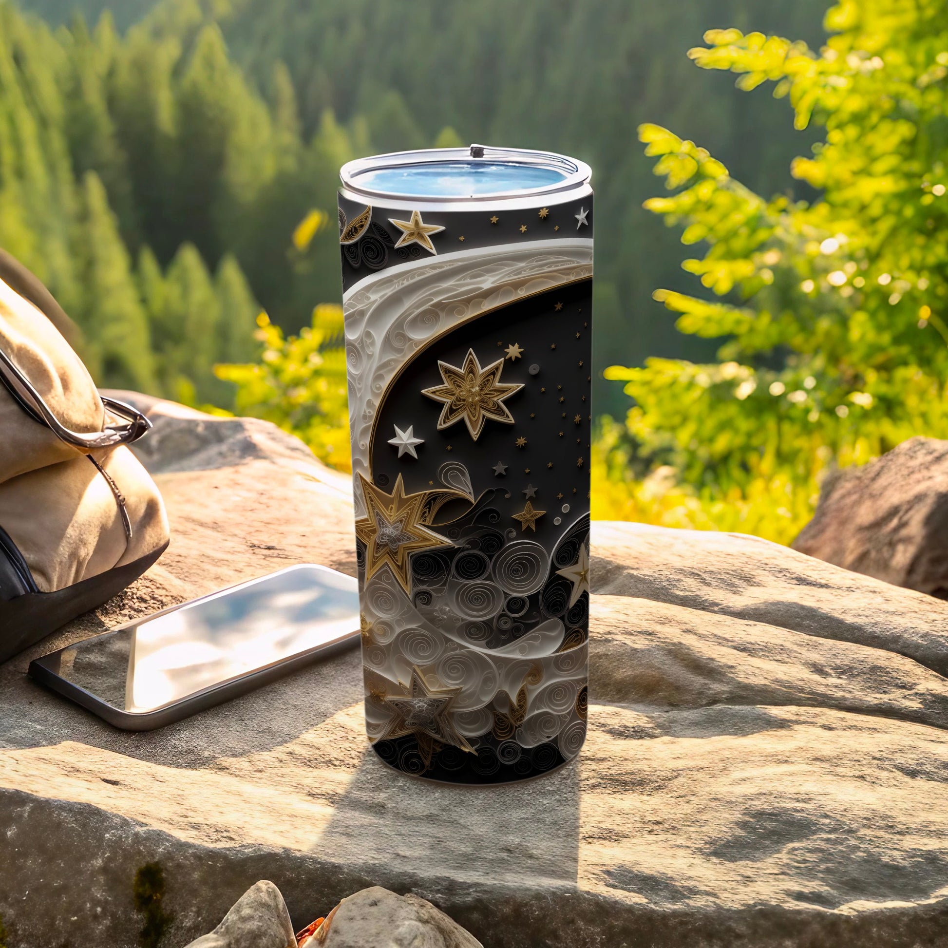 Black tumbler with gold stars and celestial swirls on a rock with a phone and bag.