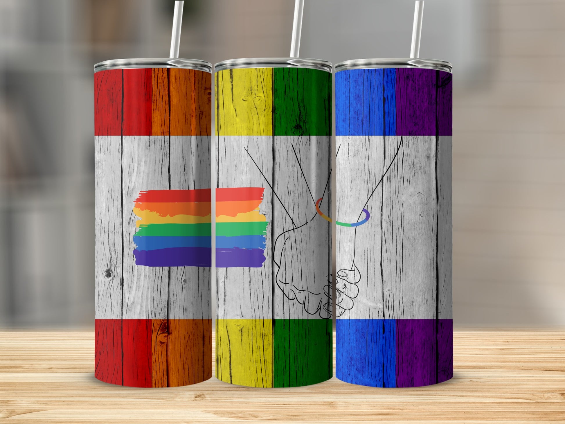 Three tumblers with rainbow flag design on wood grain background.