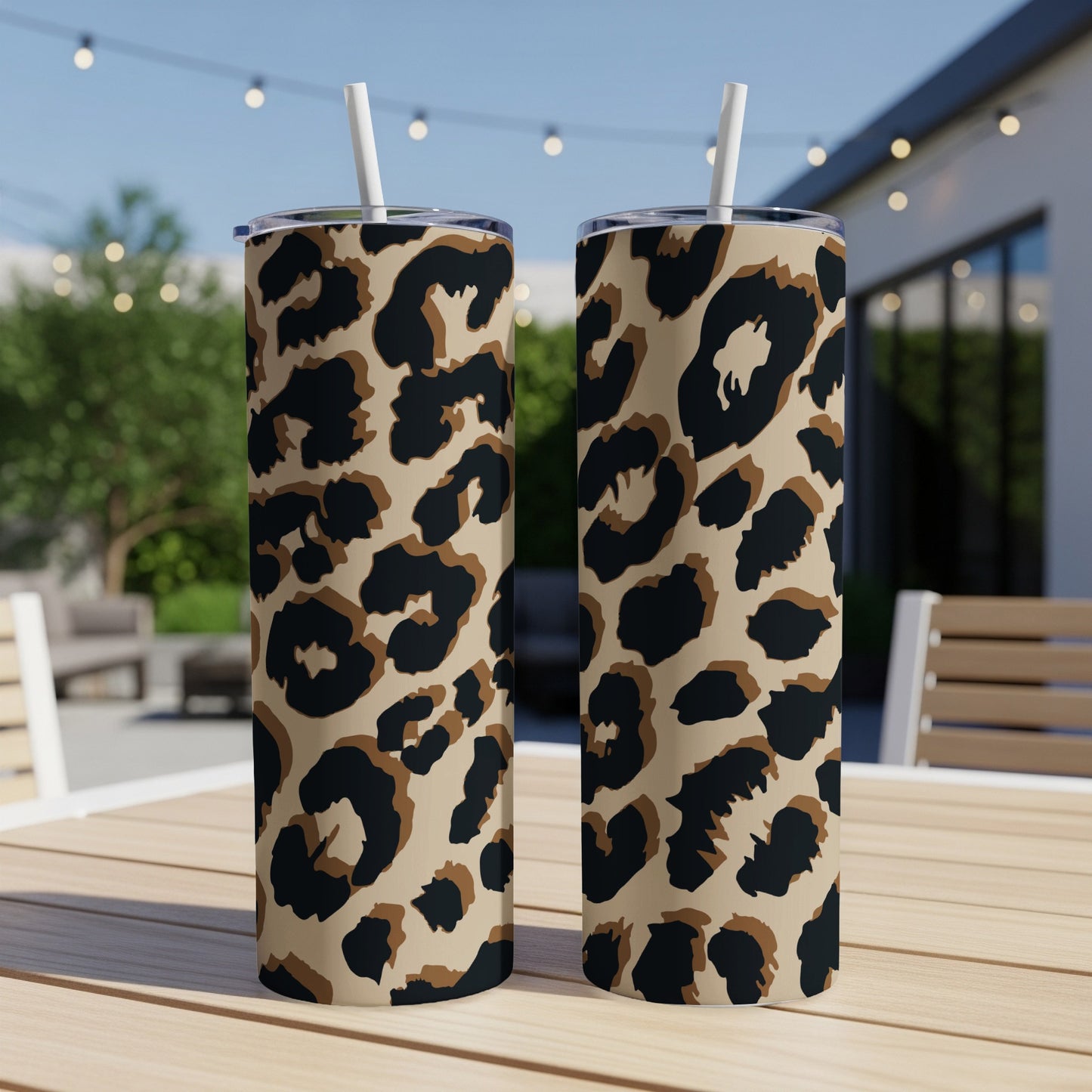 Two leopard print tumblers with white straws sit on a wooden table outdoors.