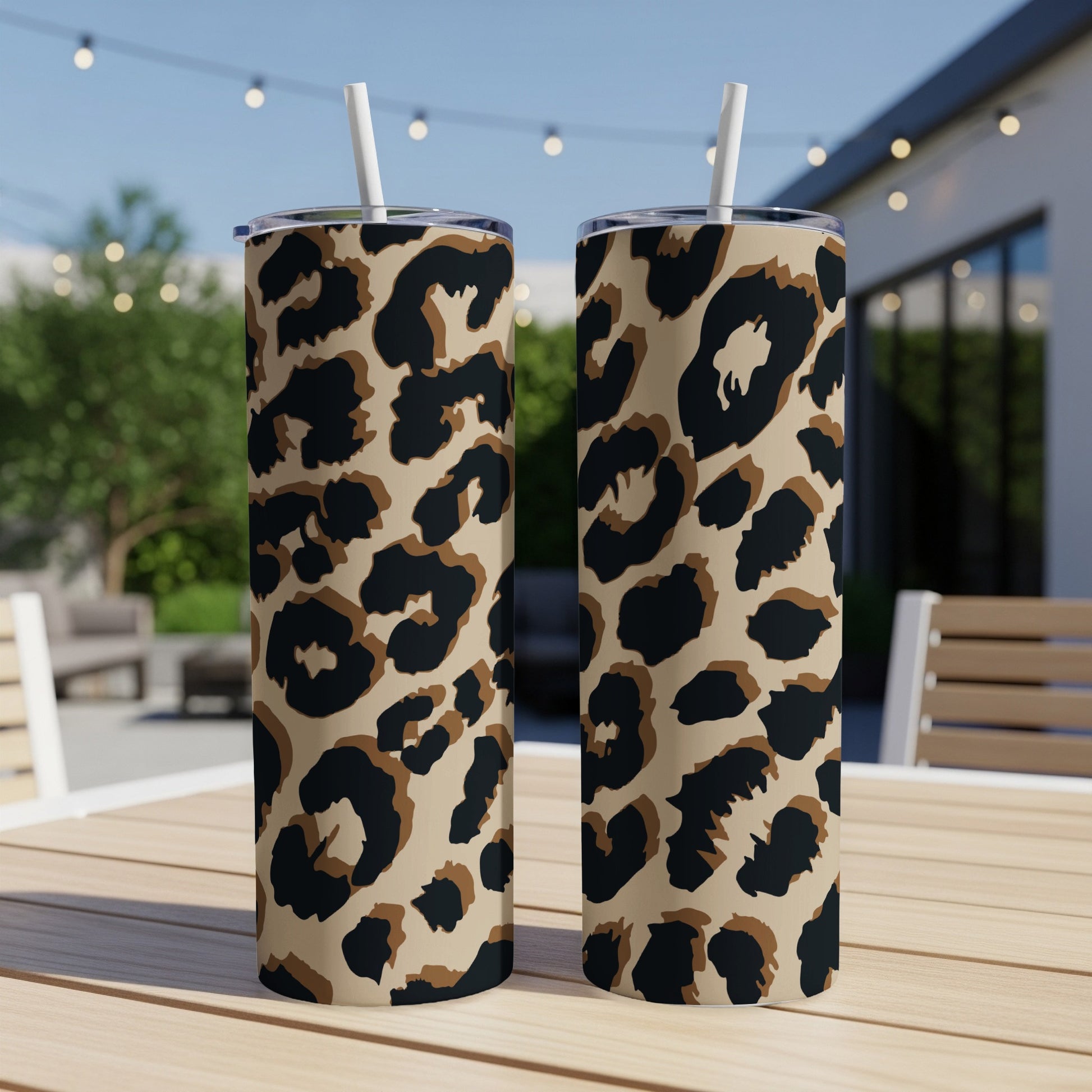 Two leopard print tumblers with white straws sit on a wooden table outdoors.