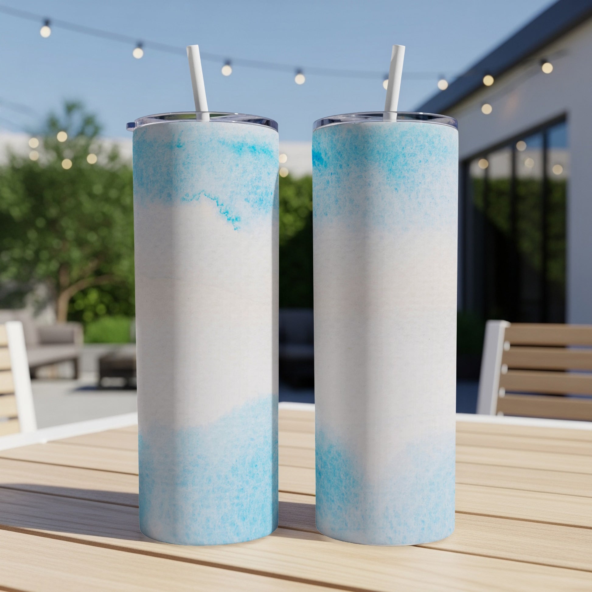 Two tall, blue-splattered tumblers with straws on a wooden table outdoors.