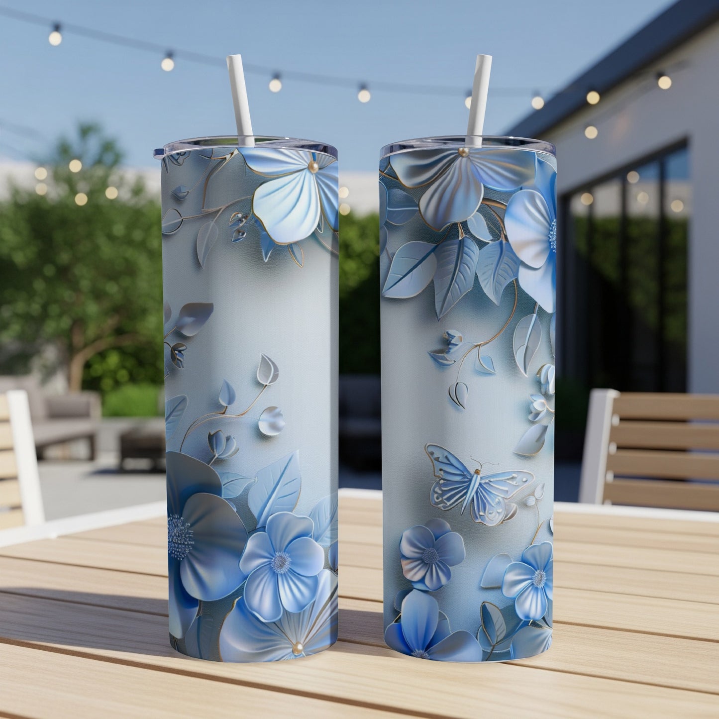 Two tall, cylindrical tumblers with a blue floral and butterfly embossed design on a patio table.