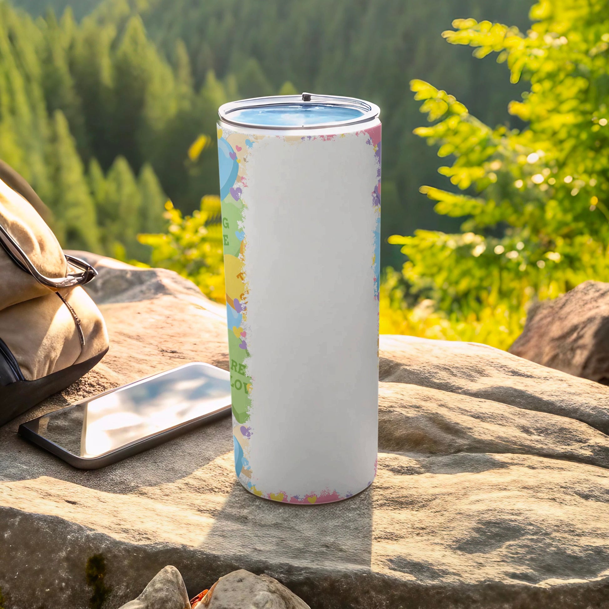 White tumbler with colorful abstract pattern and text on rocky surface with phone and bag.