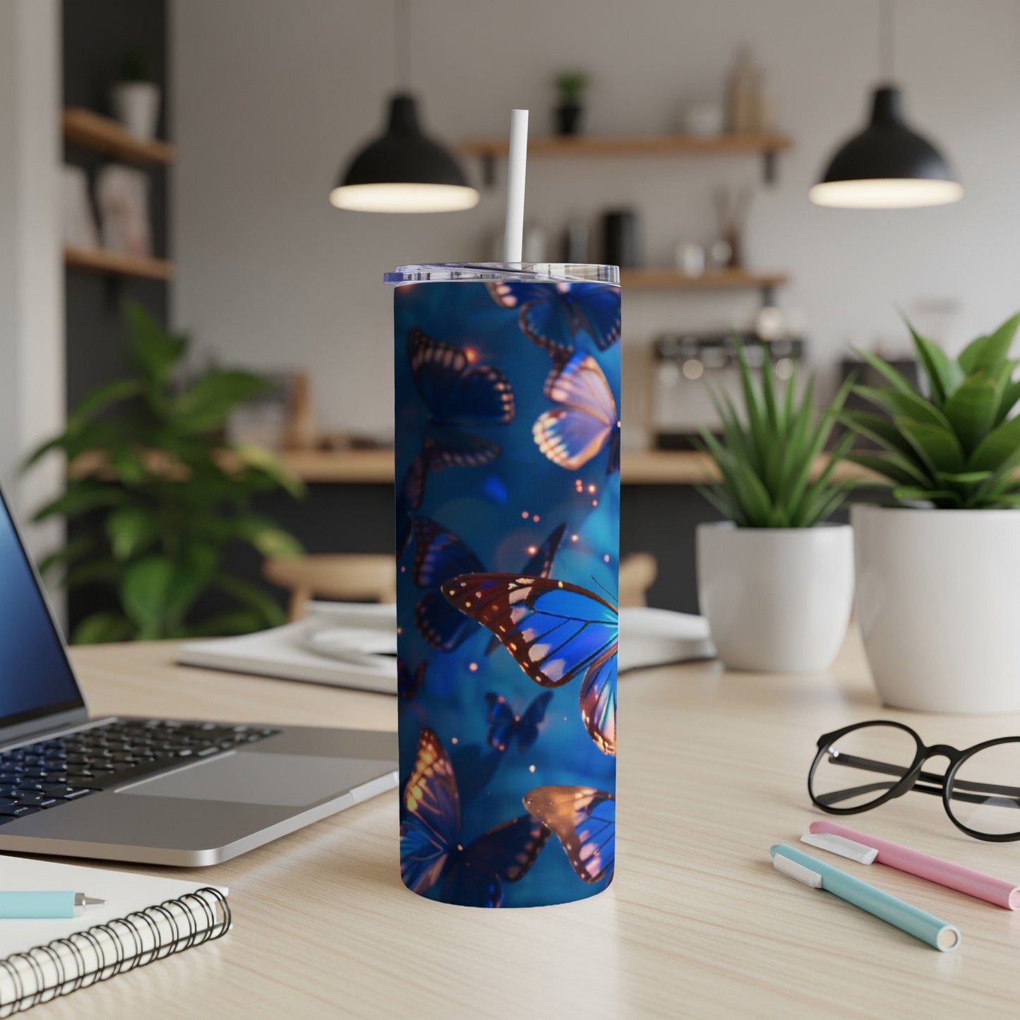 Blue butterfly tumbler with straw on desk next to laptop and pens.