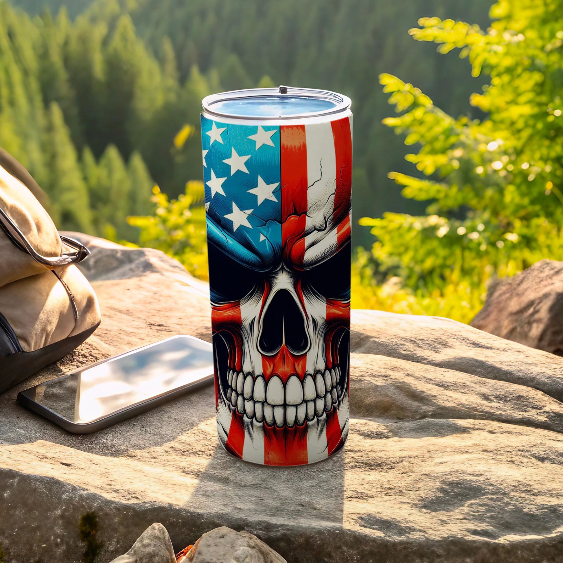 Tumbler with American flag skull design, resting on rocks with phone and backpack.