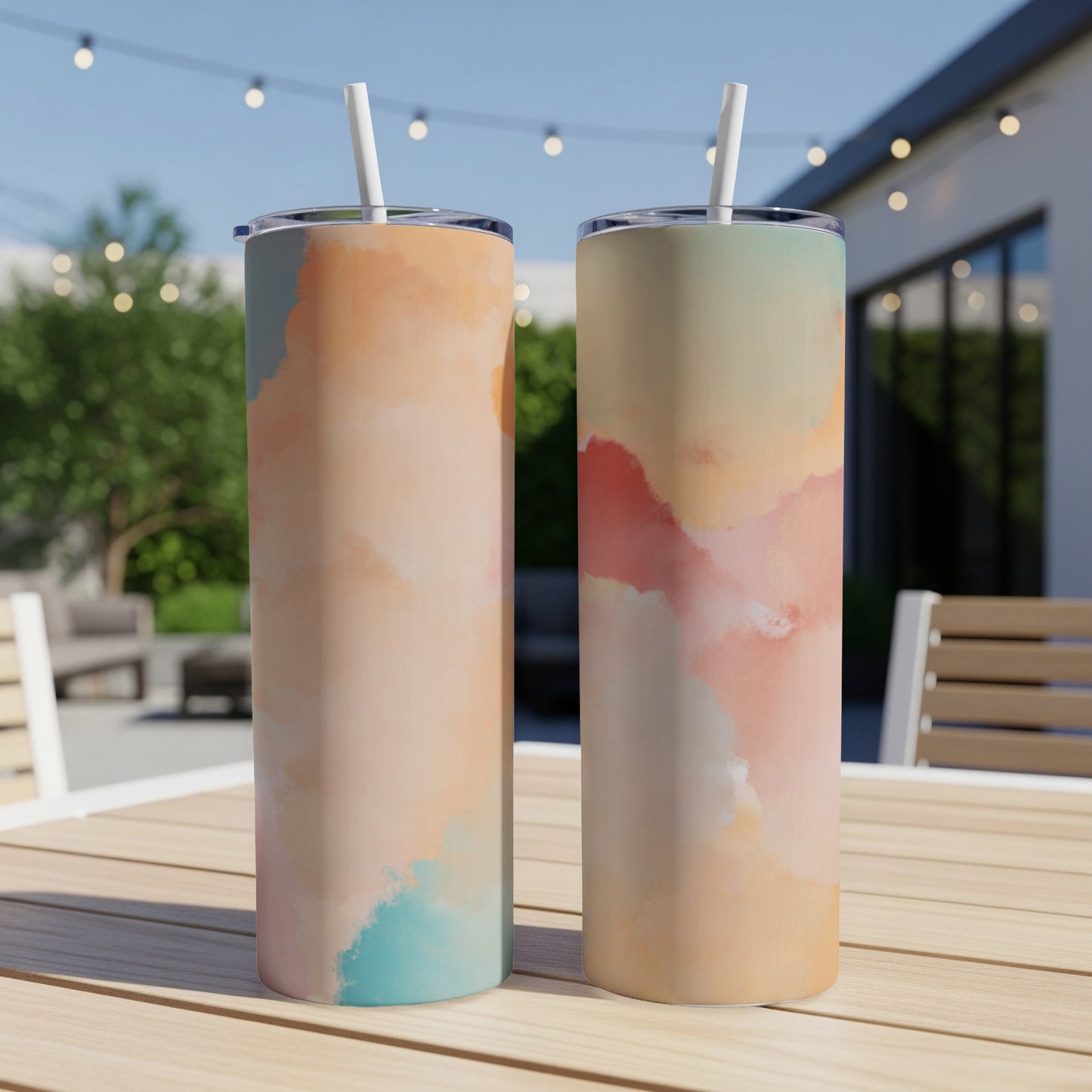 Two tumblers with pastel watercolor designs sit on a wooden table outdoors.