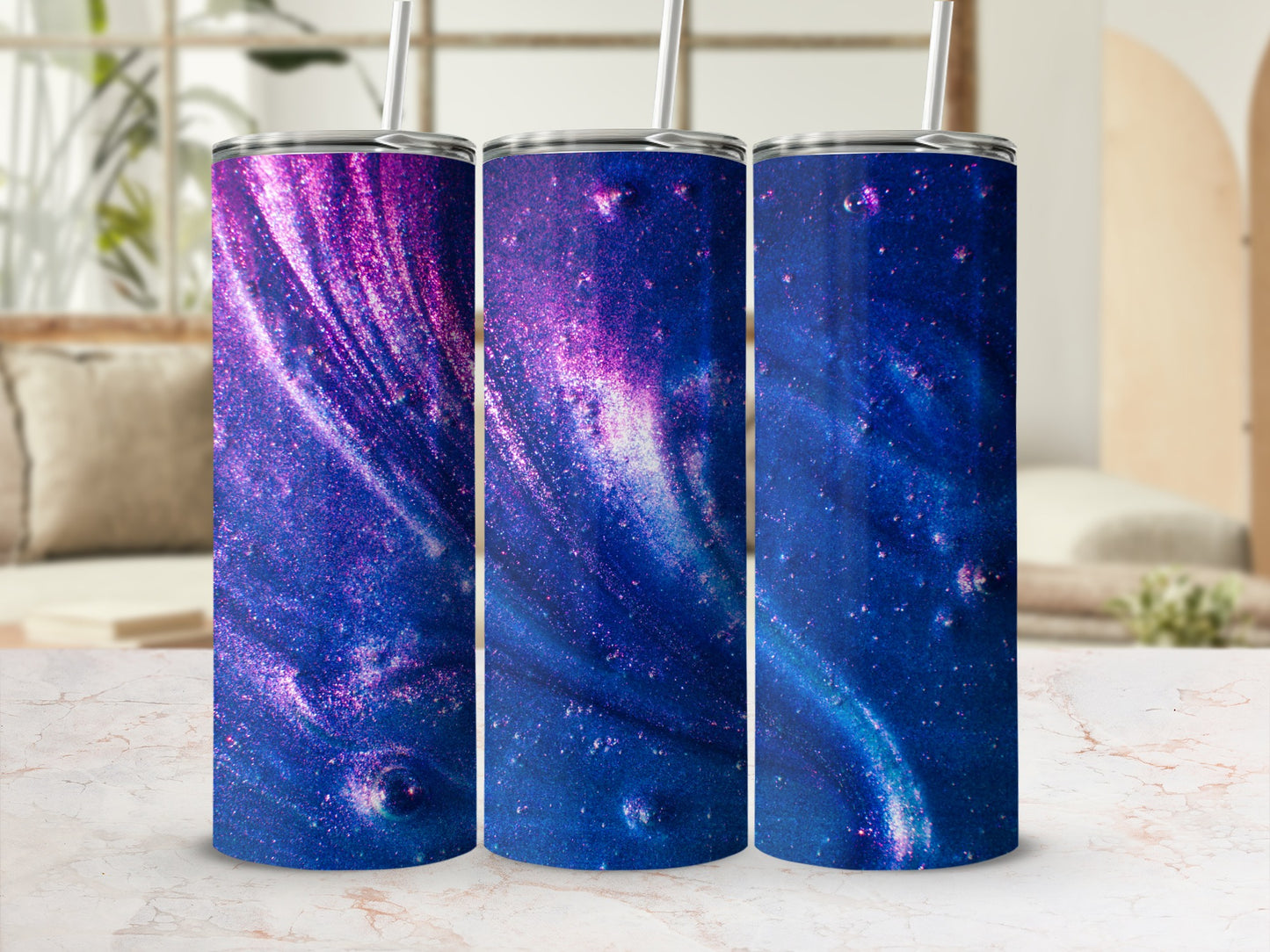 Three glittery galaxy print tumblers with straws on a marble surface.