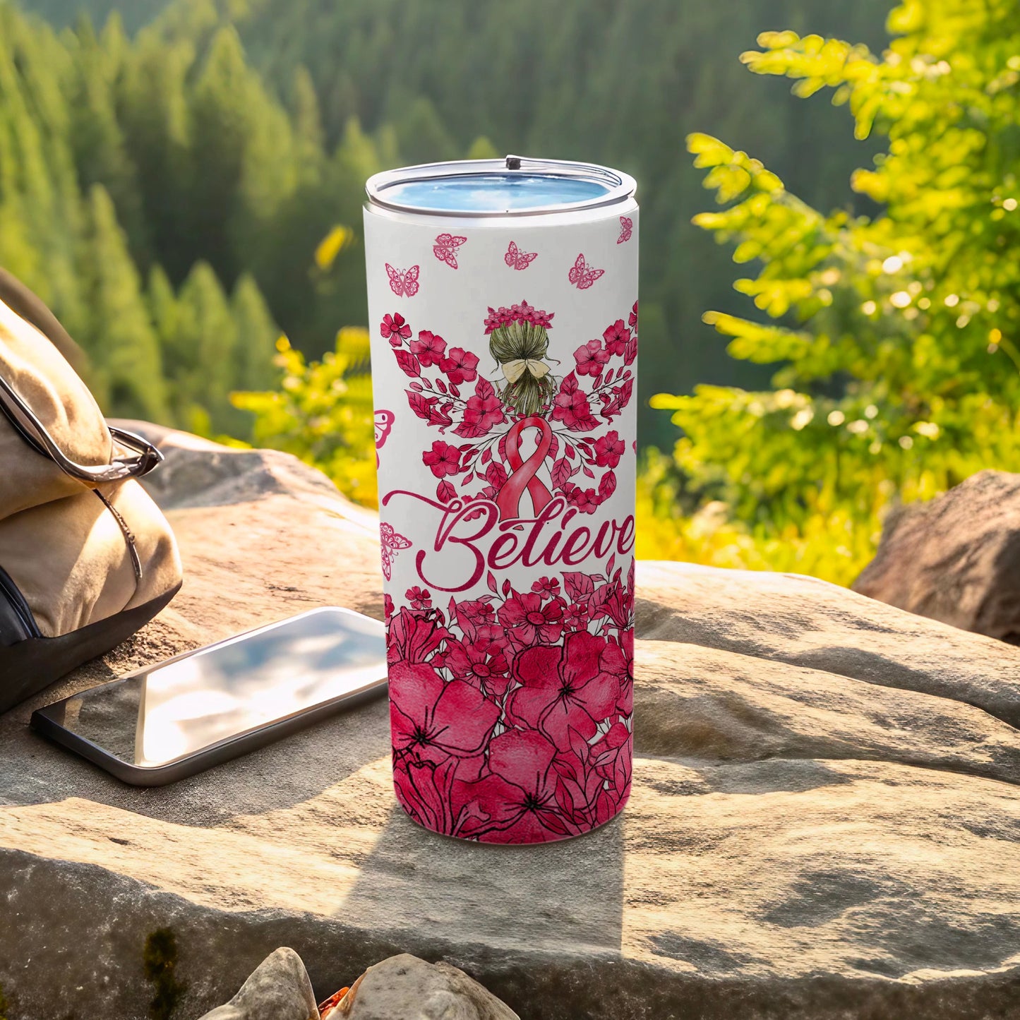 Pink floral tumbler with "Believe" text and pink ribbon, on rocks outdoors.