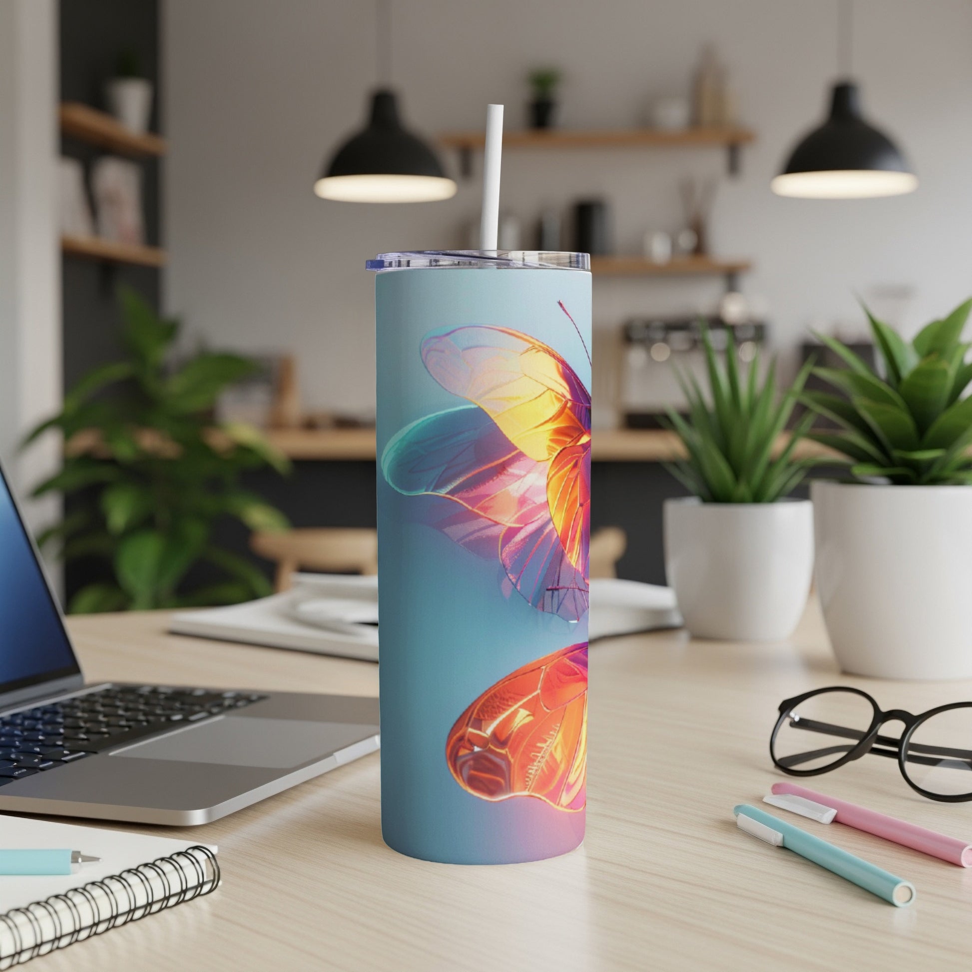 Blue tumbler with colorful butterfly design on desk next to laptop and pens.