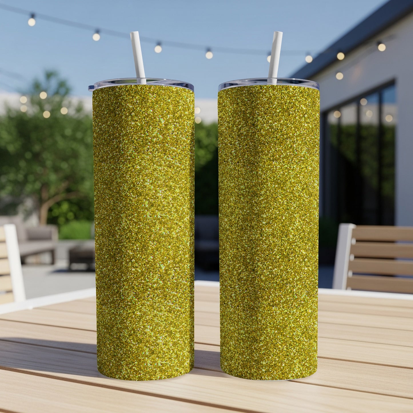 Two tall glittery gold tumblers with white straws sit on a wooden table outdoors.