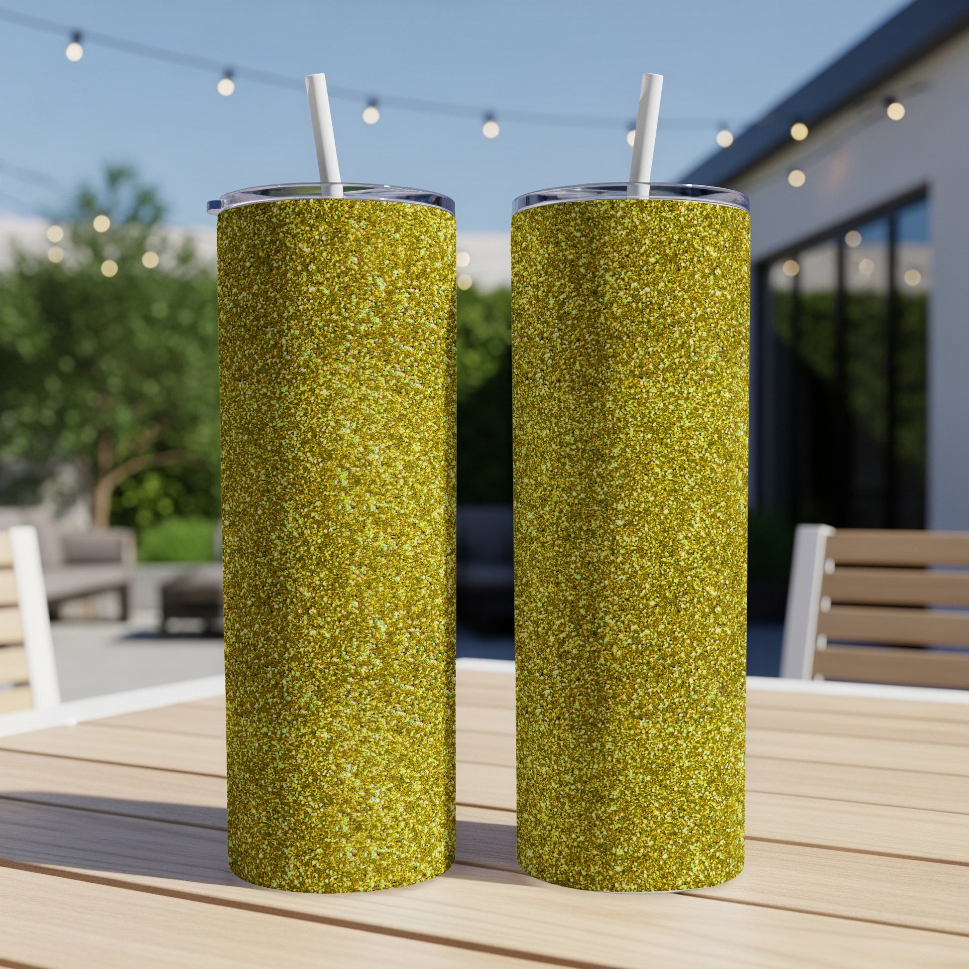 Two tall glittery gold tumblers with white straws sit on a wooden table outdoors.