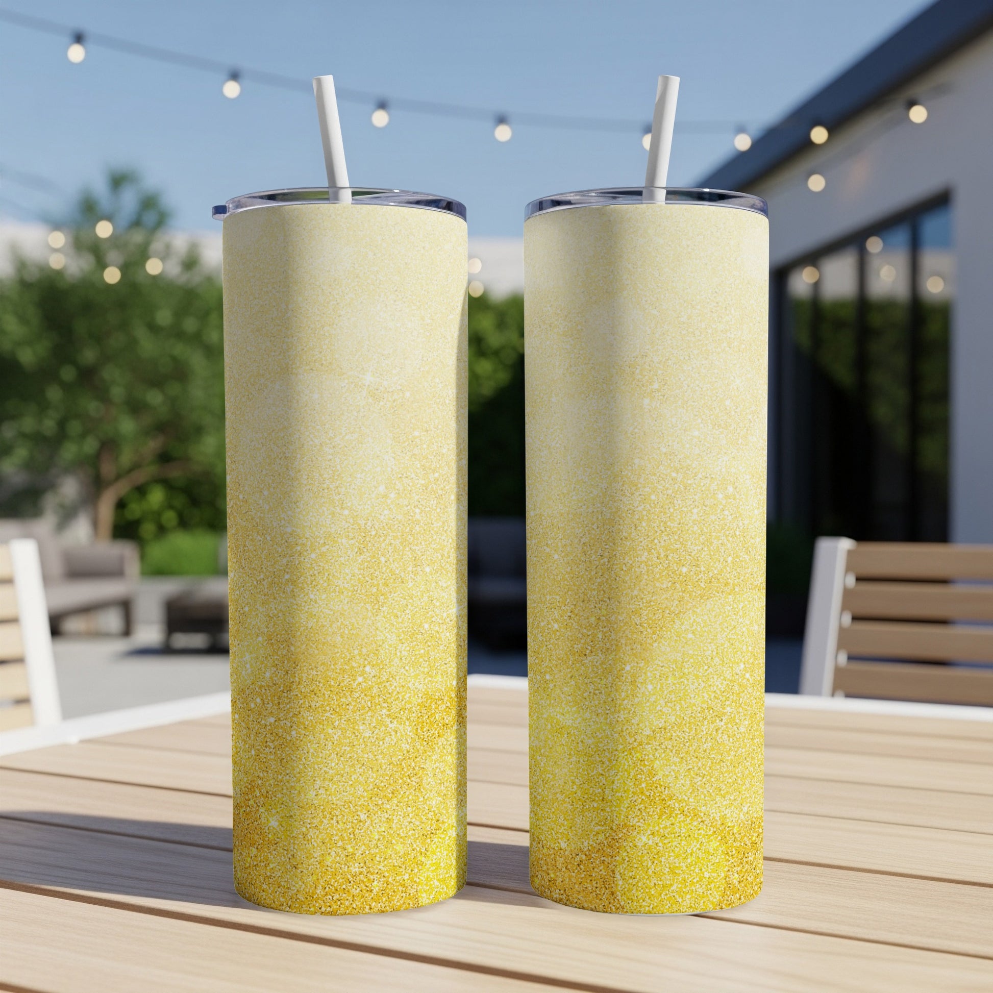 Two gold glitter tumblers with white straws stand on a wooden table outdoors.