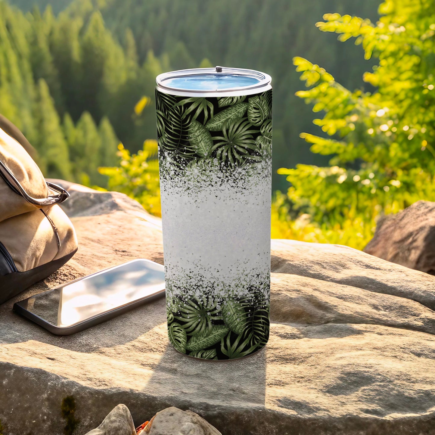 Tall tumbler with tropical leaf pattern on rocky outdoor surface near backpack and phone.