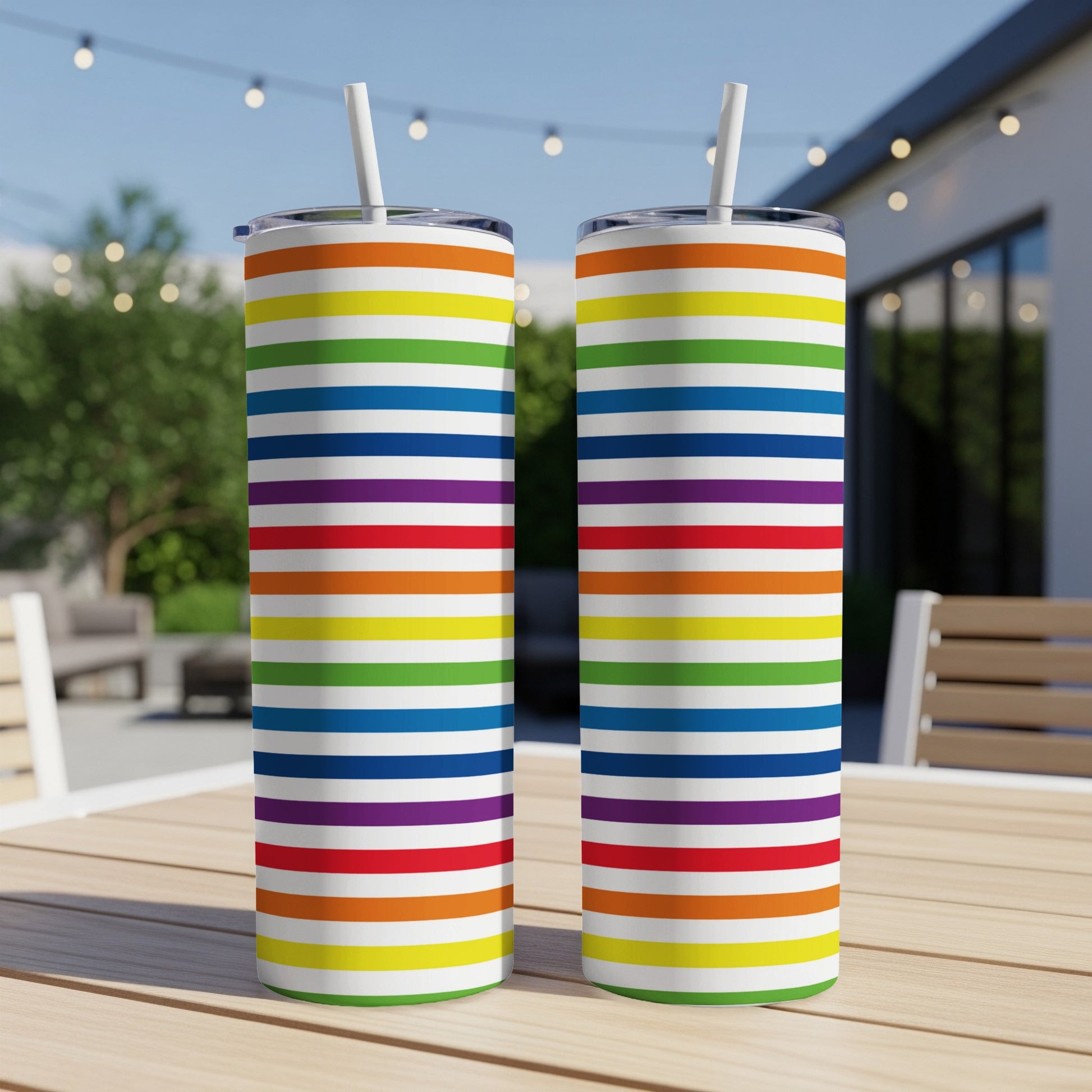 Two rainbow striped tumblers with straws on a wooden table outdoors.