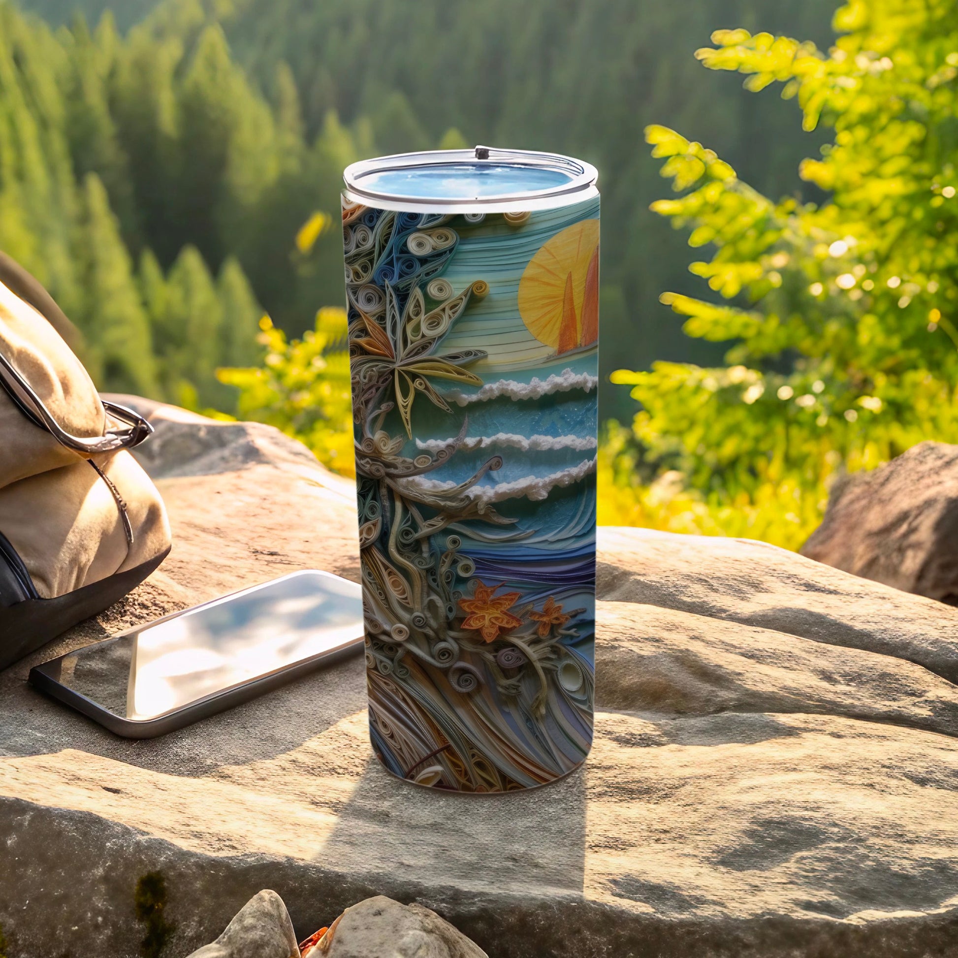 Tumbler with ocean waves and sun design, resting on rock with phone and bag.