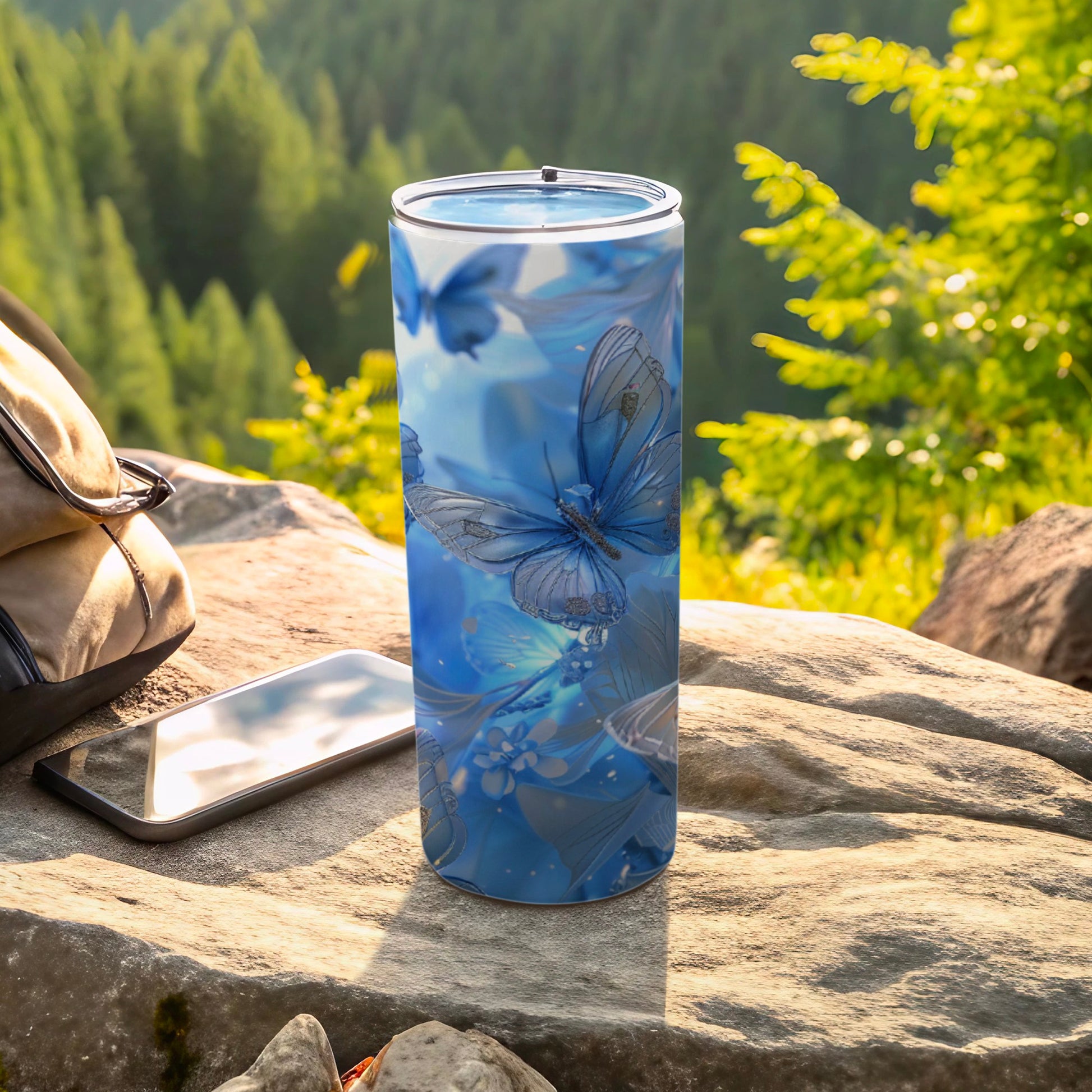 Blue butterfly-patterned tumbler on rock with phone and backpack.
