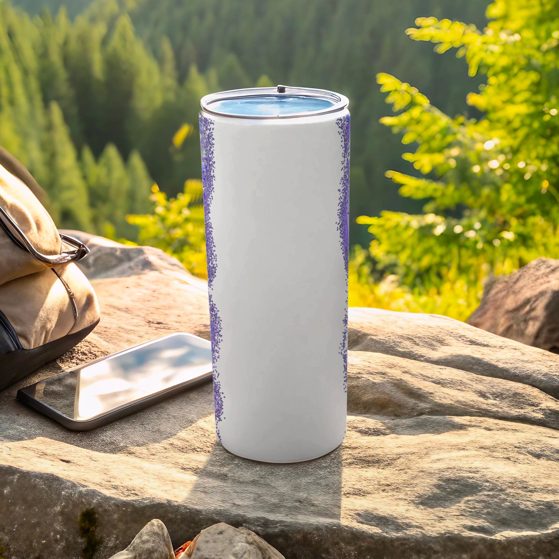 White tumbler with purple speckled pattern on rock with phone and backpack.
