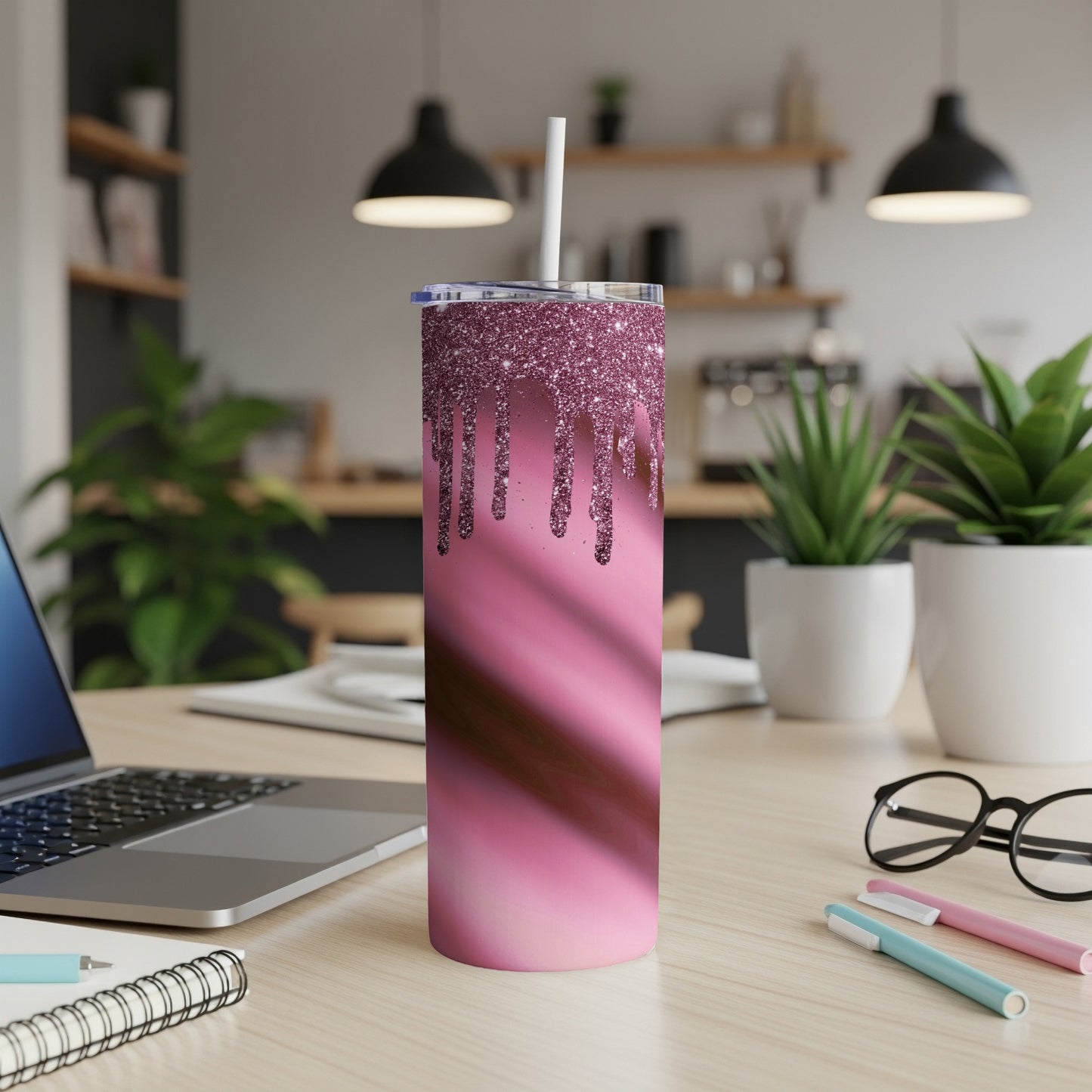 Pink glitter dripping tumbler with straw on a desk beside laptop and glasses.